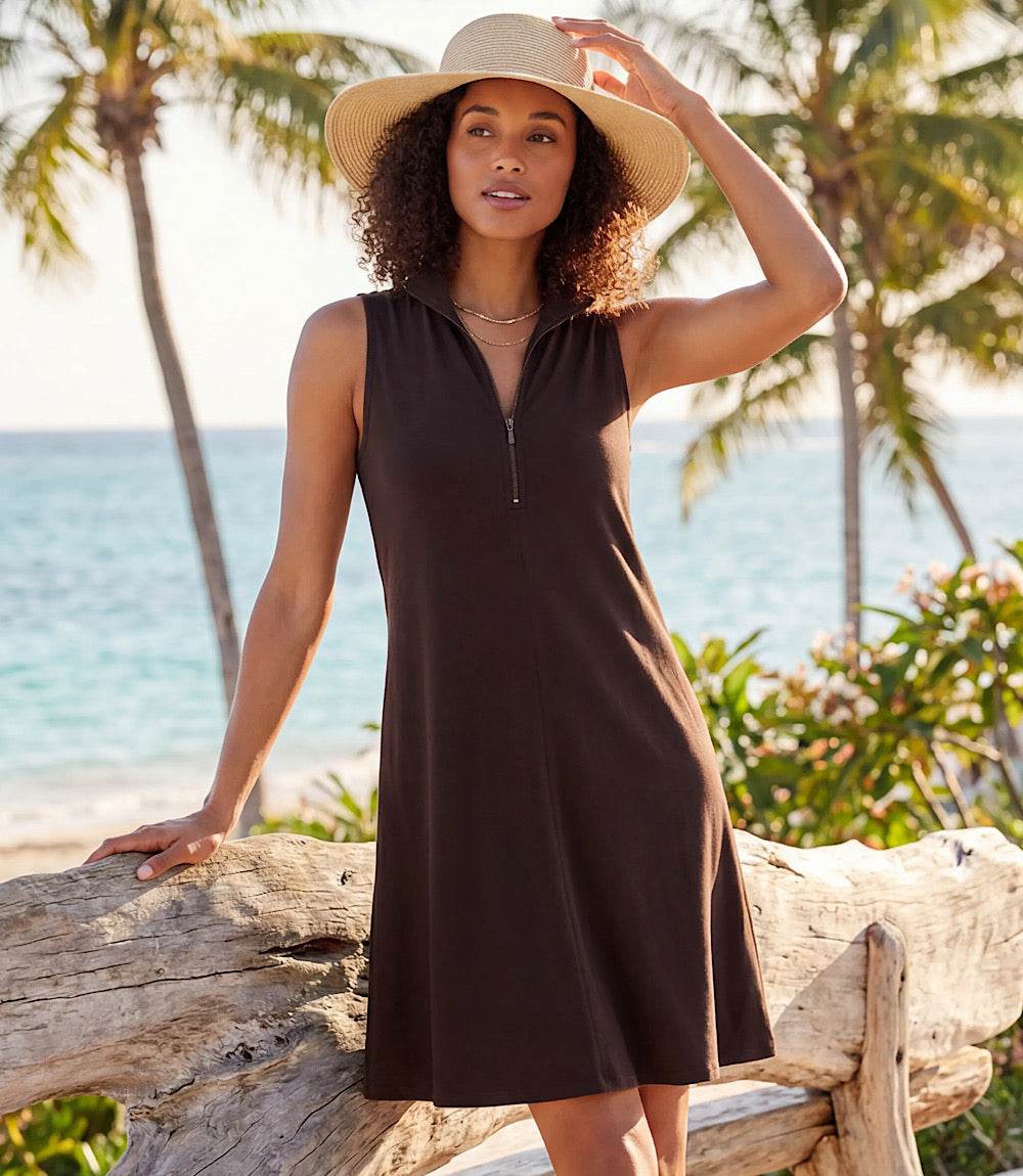 A woman wearing a sleeveless dark brown dress with a half-zip neckline and a wide-brim straw hat, posing on a beach with palm trees and the ocean in the background.