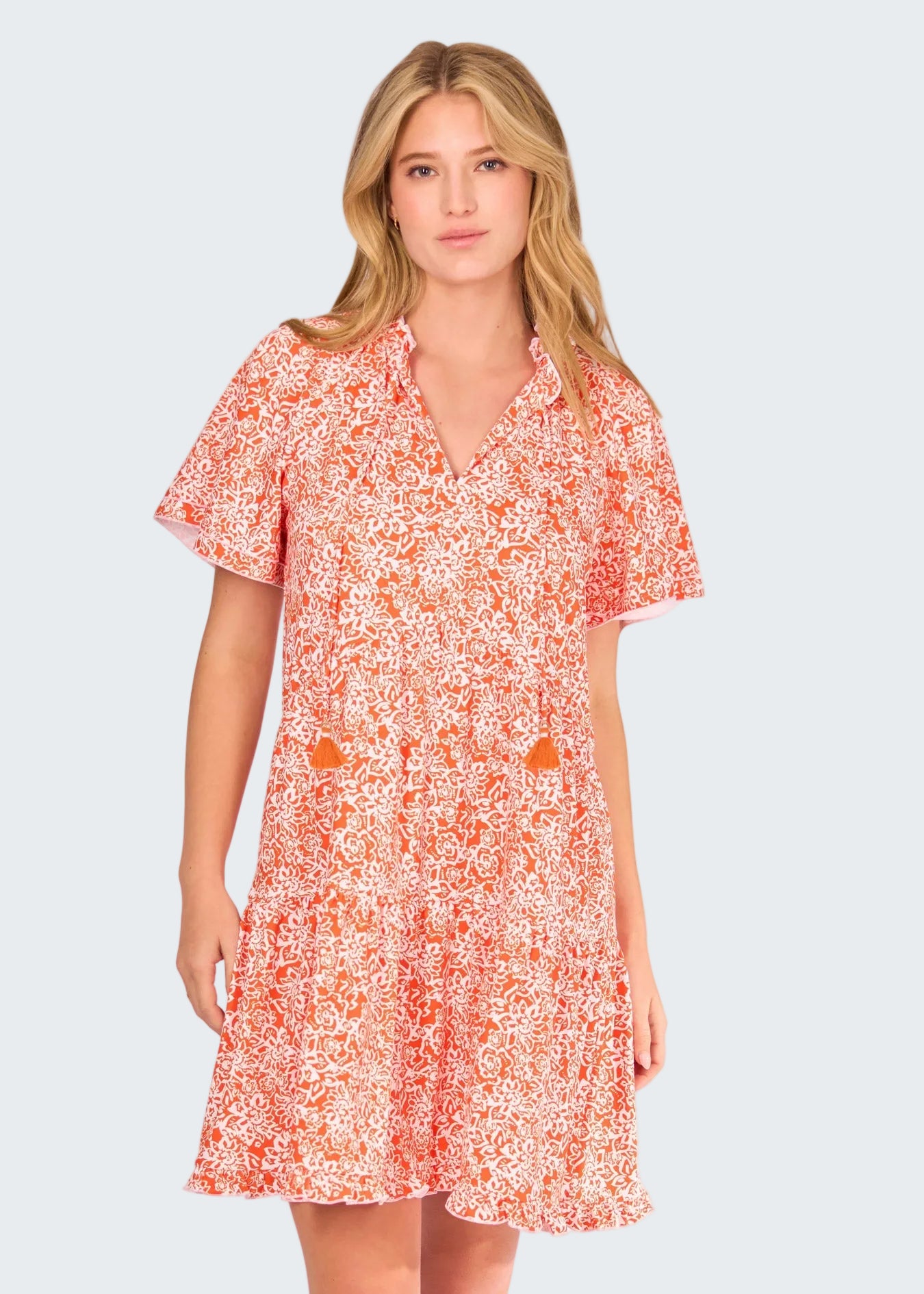 A woman modeling a short-sleeve V-neck dress with a dense orange and white floral print against a solid white background.