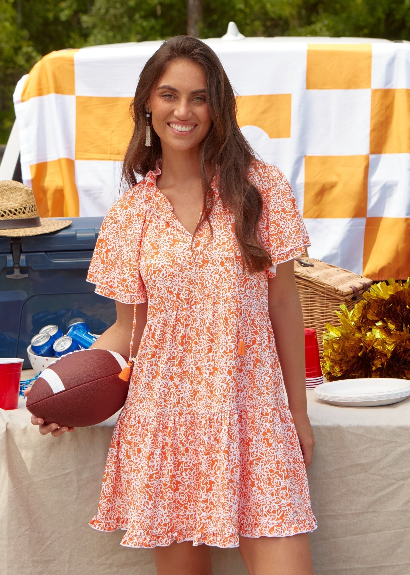 Woman in a floral dress holding a football at a picnic table with a checkered tablecloth.