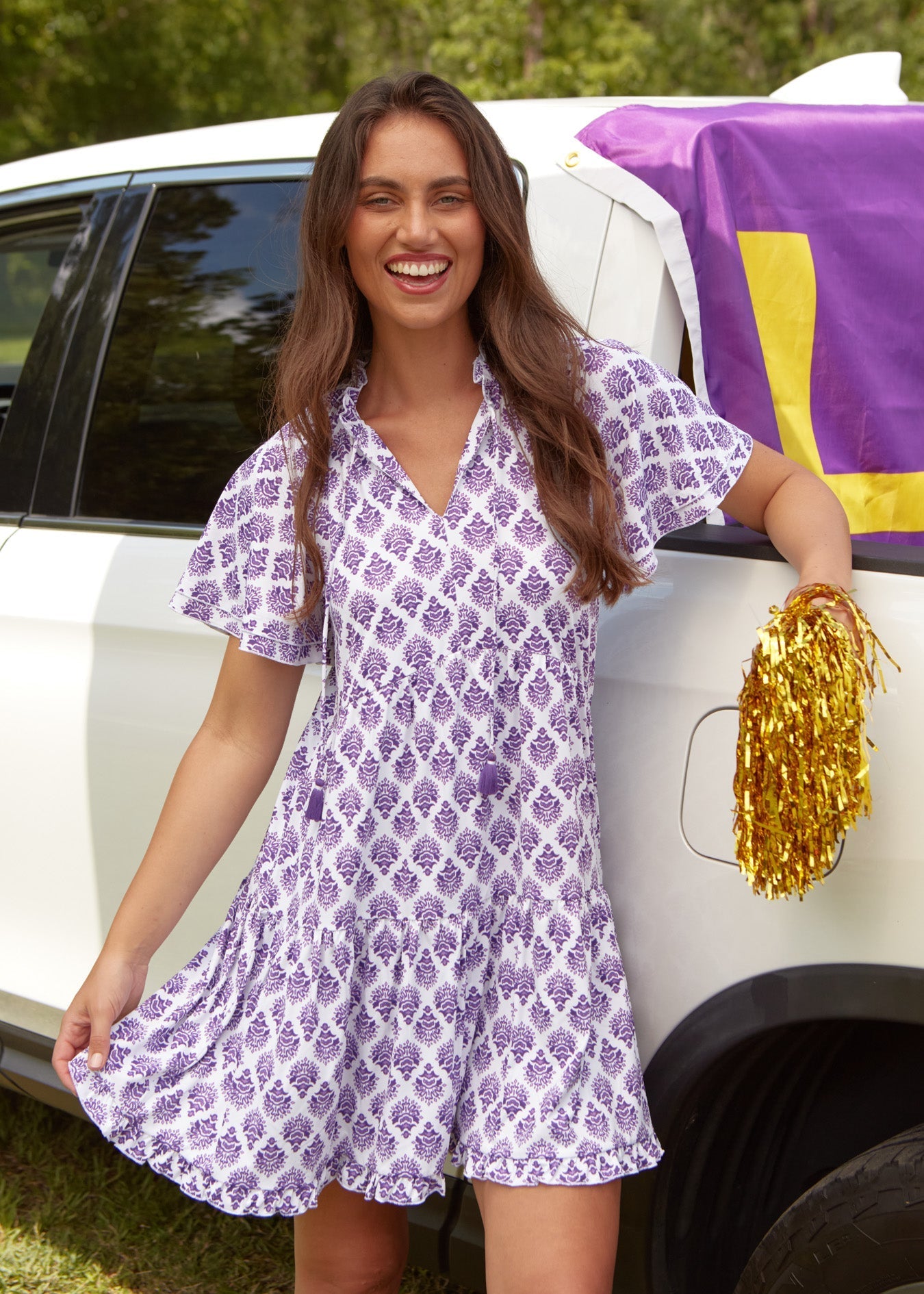 Woman in a purple and white dress holding gold pom-poms next to a white car with a purple and yellow cover.