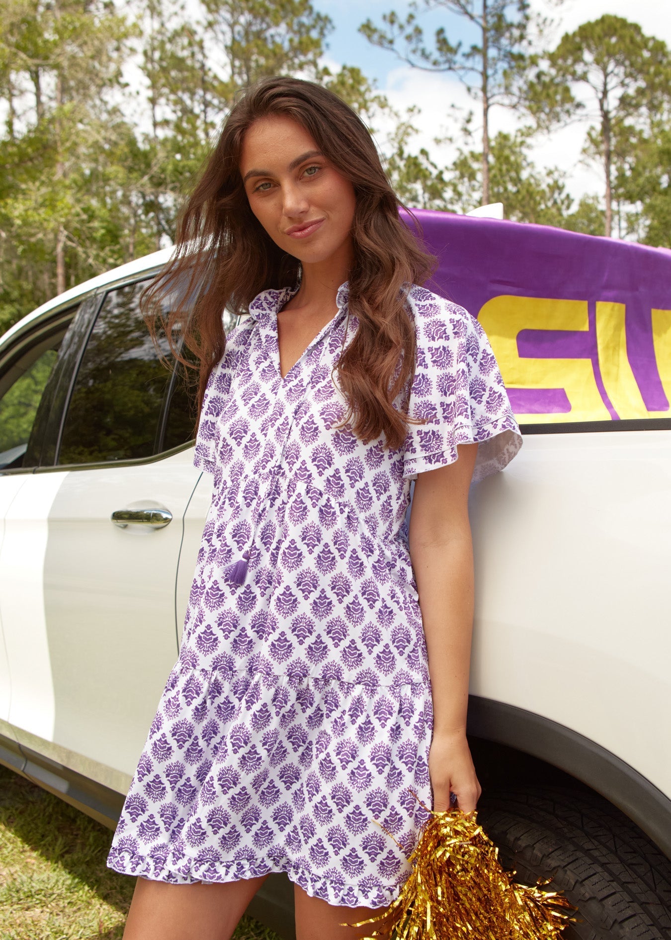 Woman in a purple patterned dress standing next to a white car with a purple surfboard.