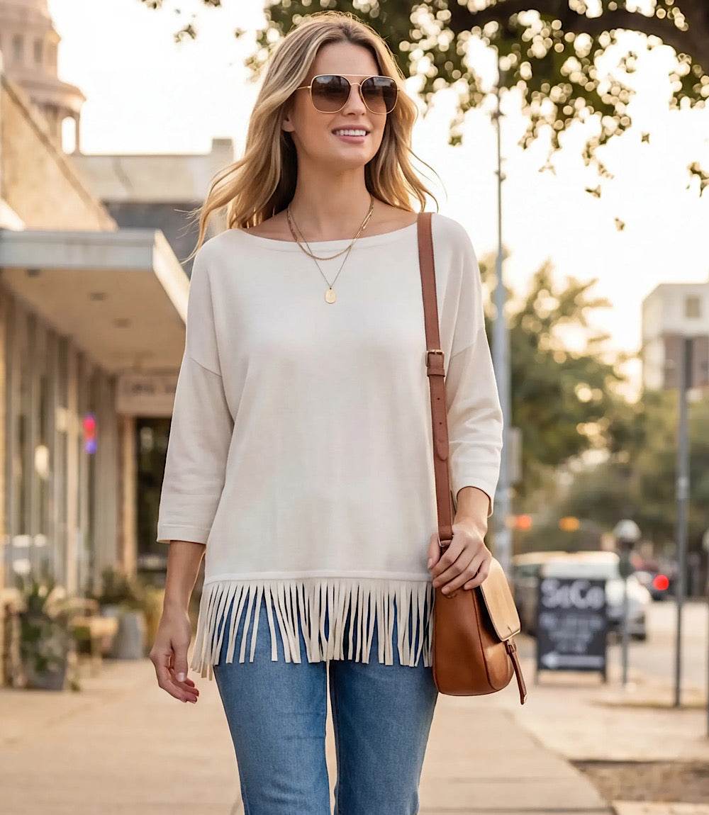 A woman walking on a city sidewalk wearing a cream-colored, three-quarter sleeve fringe top, blue jeans, sunglasses, and carrying a brown leather shoulder bag.