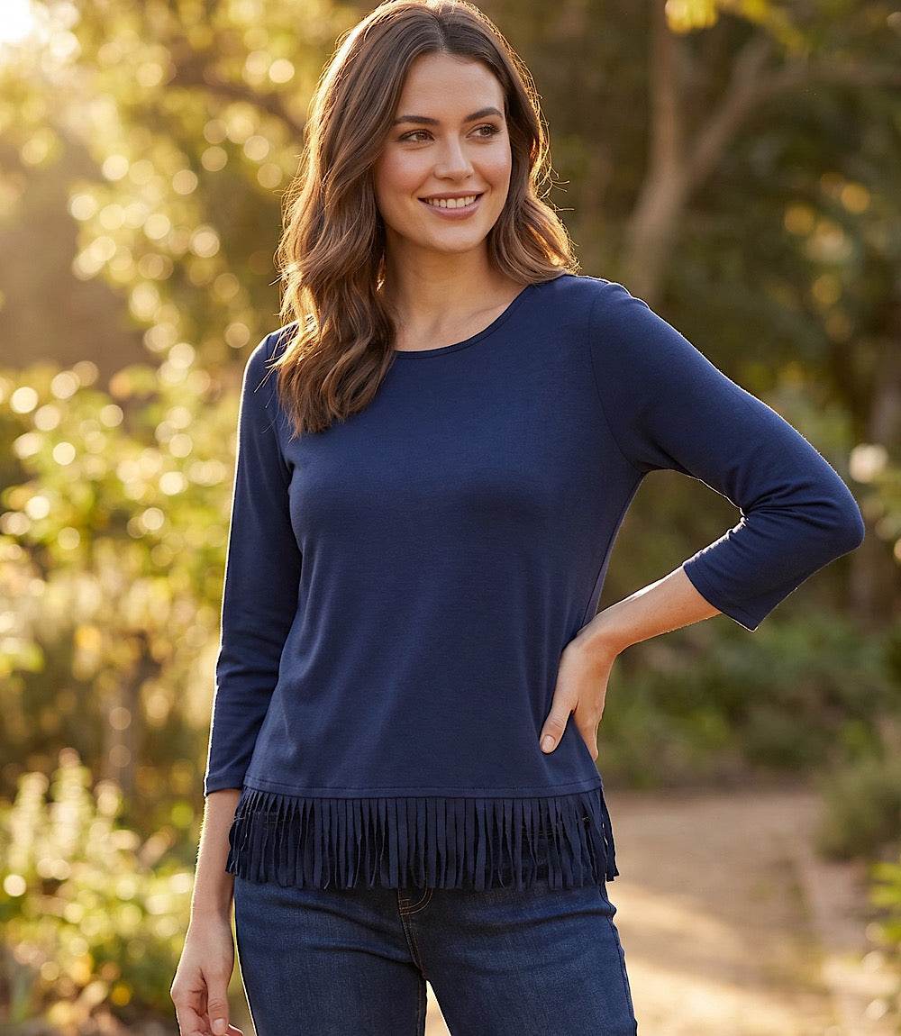 A woman wearing a navy blue long-sleeve top with fringe trim at the hem, paired with dark blue jeans, standing outdoors in a sunlit garden setting.