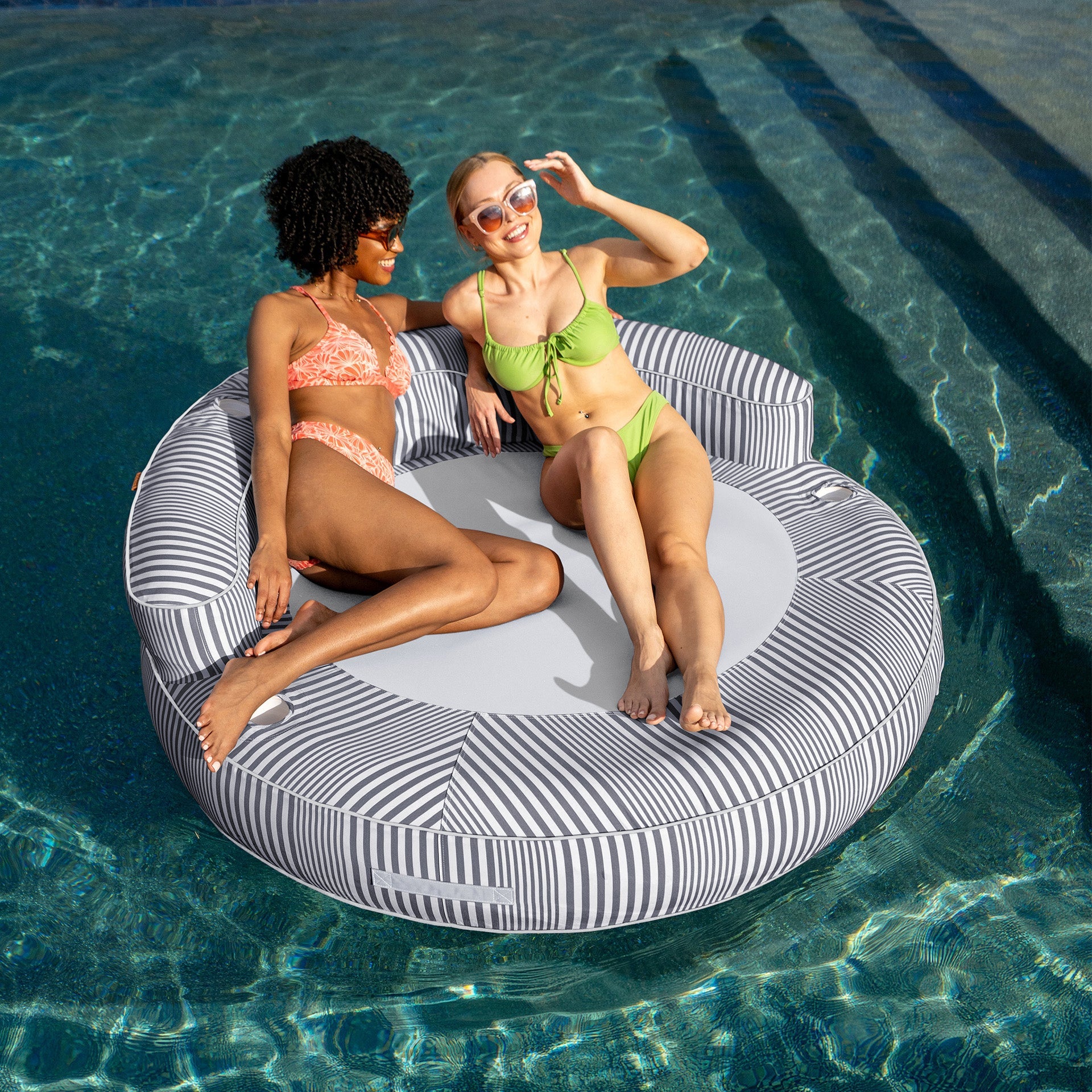 Two women in swimsuits sitting on a circular pool float in a pool.