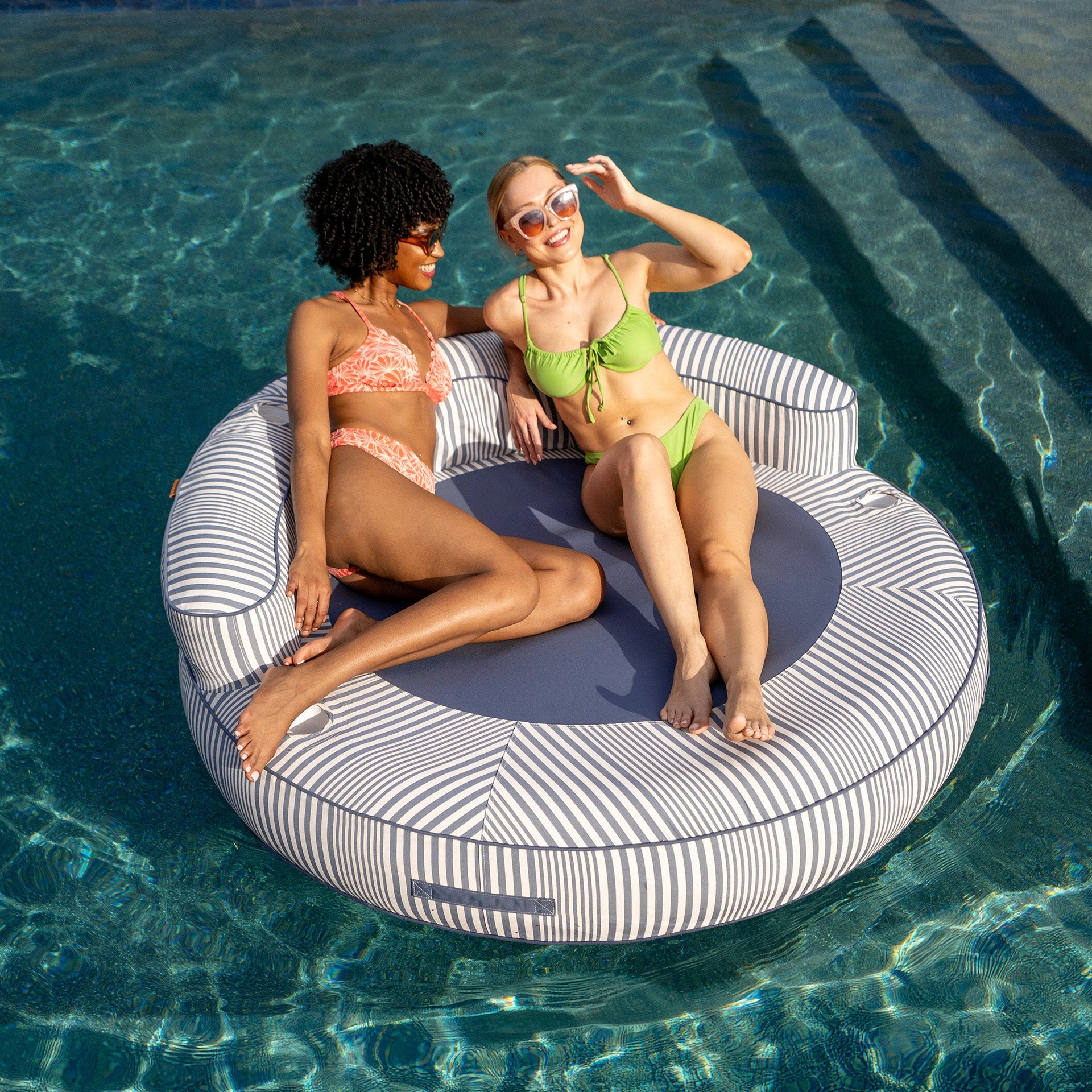 Two women sitting on a striped pool float in a pool