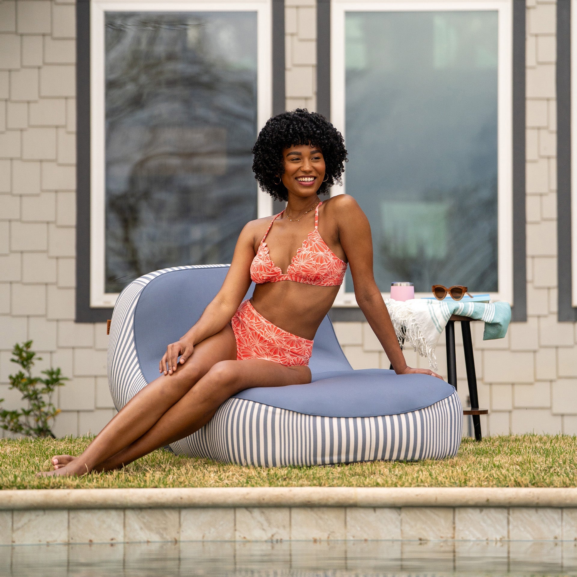 Woman in a pink bikini sitting on a striped bean bag by a poolside.