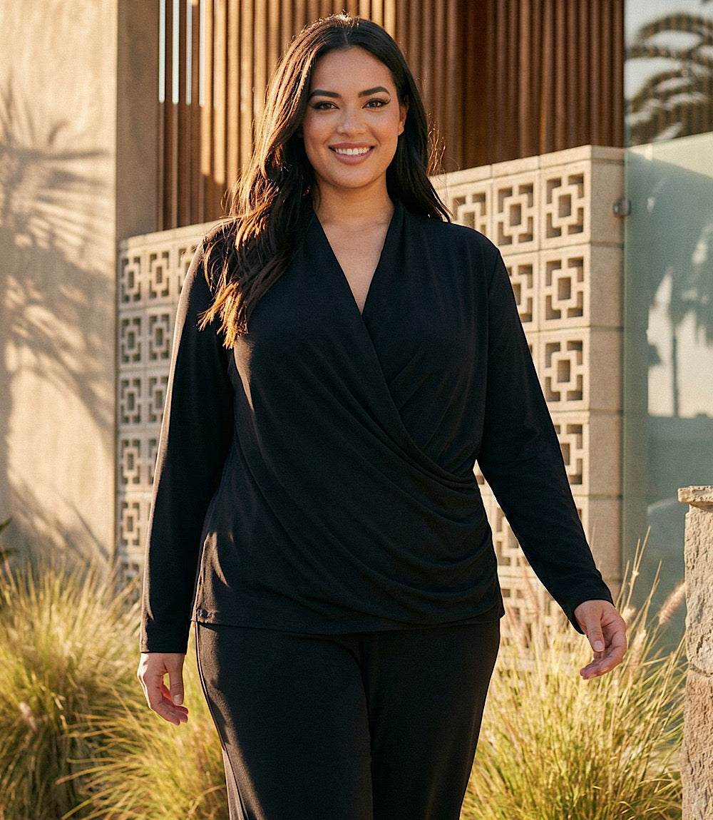 A woman wearing a black long-sleeve wrap-style top and matching black pants, standing outdoors in front of a modern building with geometric wall panels and grass.