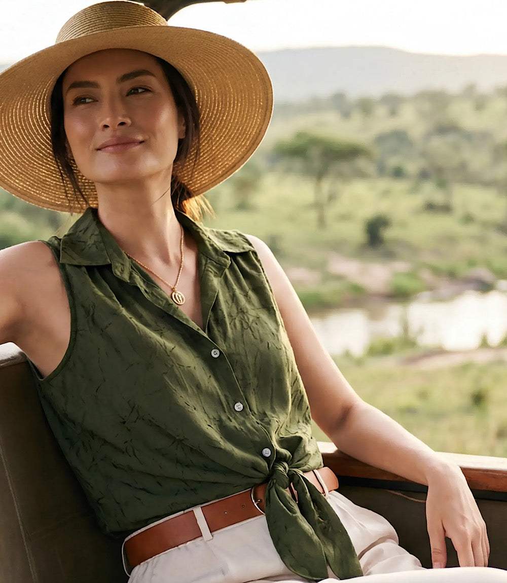 A woman wearing a wide-brimmed straw hat, an olive green sleeveless button-up shirt tied at the waist, and white trousers, seated in a safari-style vehicle with a savanna landscape in the background.