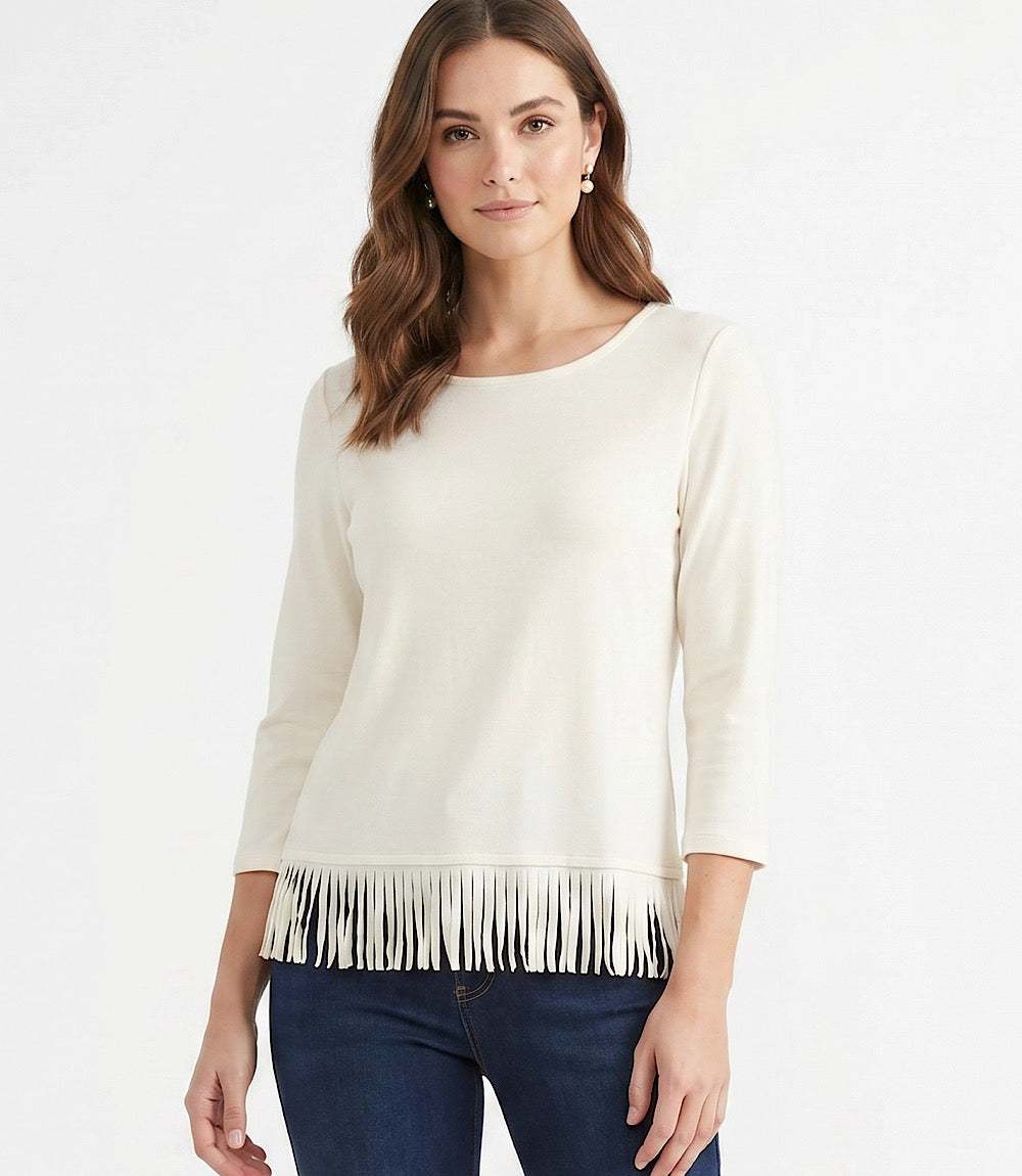 A woman wearing a cream-colored three-quarter sleeve top with fringe trim at the hem, paired with dark blue jeans, against a plain white background.