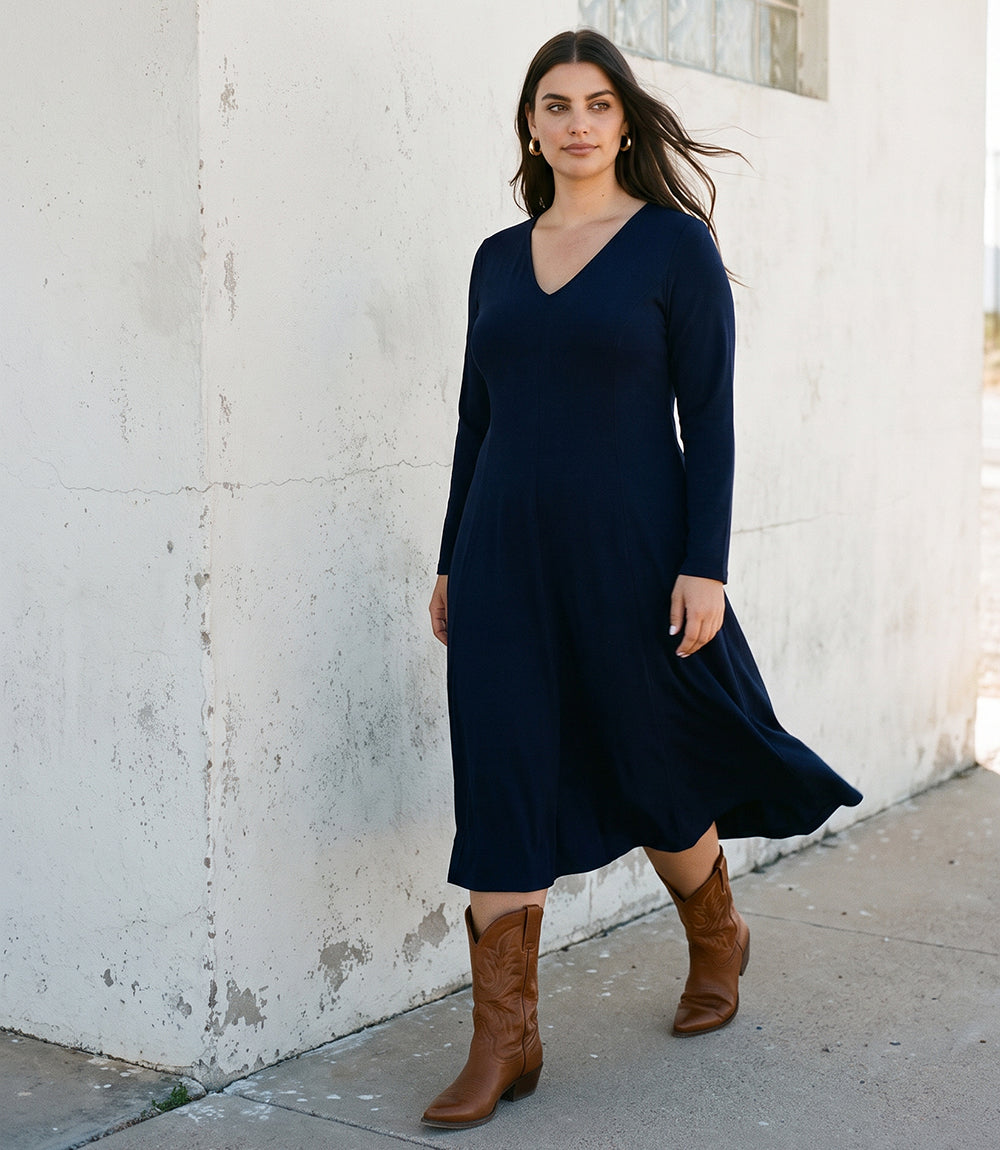 A woman wearing a navy blue long-sleeve midi dress and brown leather boots, standing outdoors against a white concrete wall.
