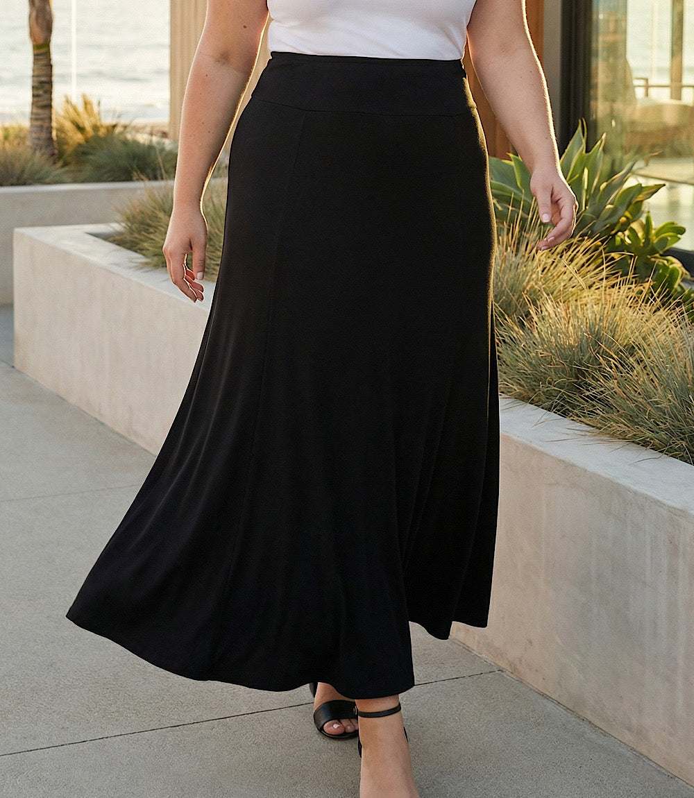 A woman wearing a long black maxi skirt with a white top, standing outdoors on a concrete walkway next to a planter bed.