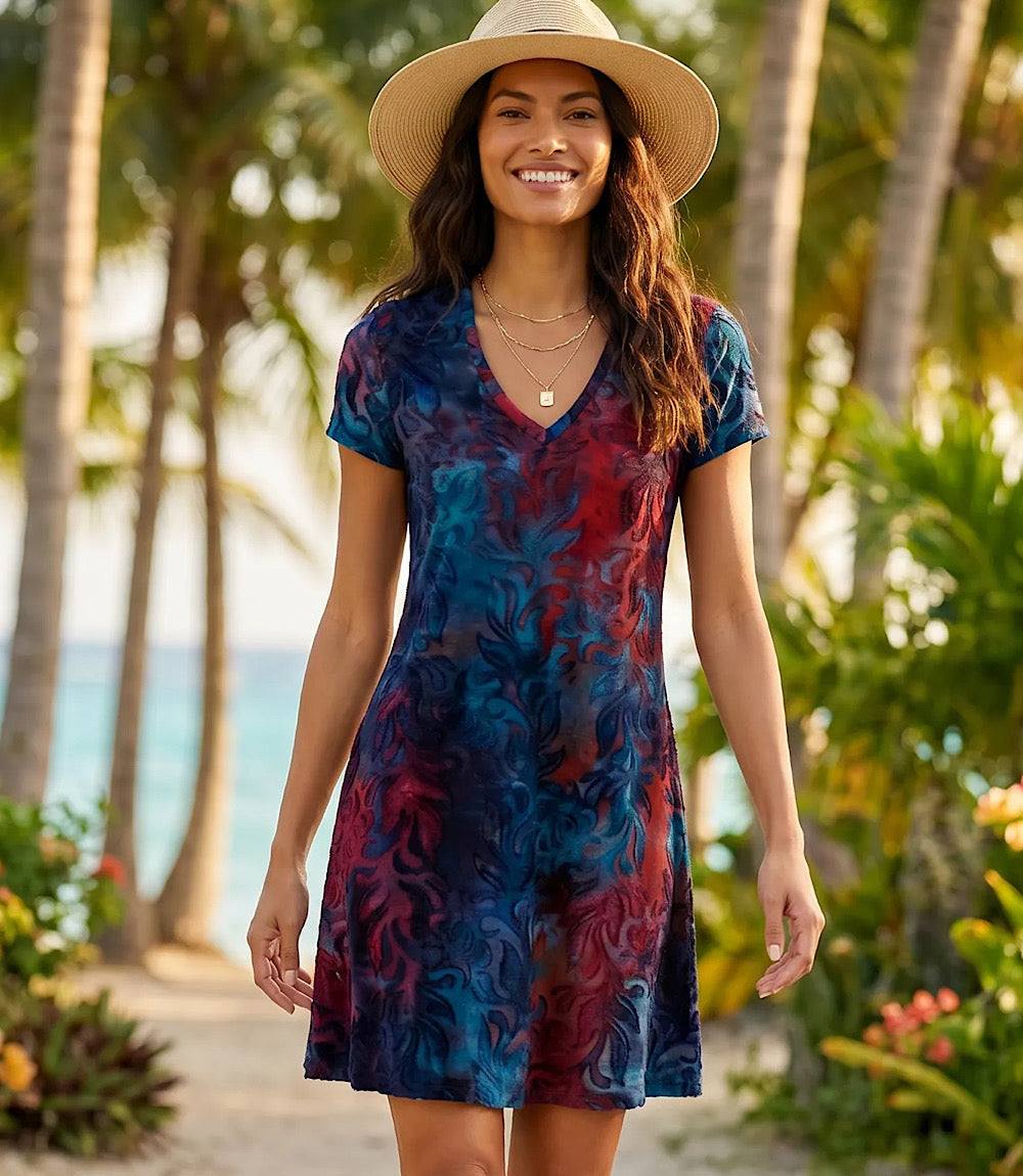 A woman wearing a short-sleeve V-neck dress with a blue, red, and purple abstract swirl pattern, a beige wide-brim hat, and layered gold necklaces, standing outdoors in front of a tropical beach background with palm trees.