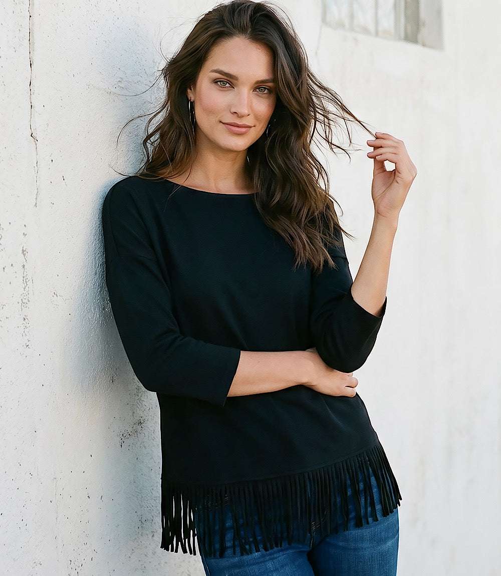 A woman with long brown hair leans against a white textured wall, wearing a black three-quarter sleeve top with fringe trim at the hem and blue jeans.