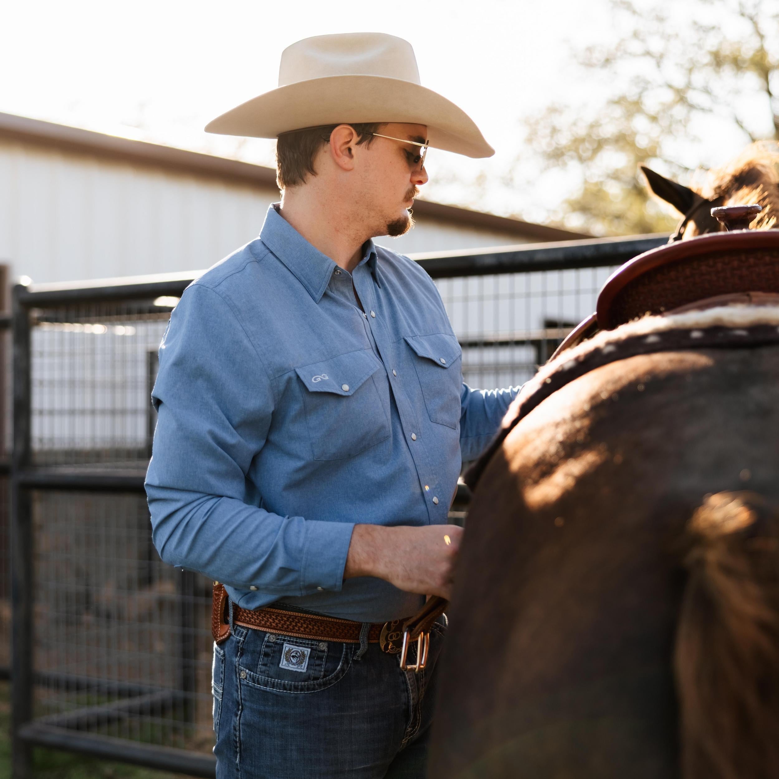 Man in a cowboy hat and blue shirt standing next to a horse, adjusting the saddle.