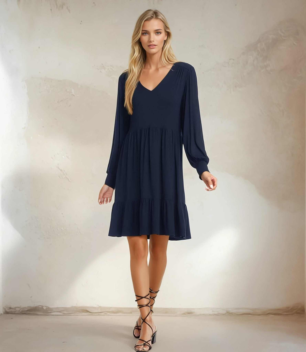 A woman modeling a navy blue long-sleeve V-neck mini dress with a gathered waist and flared ruffle hem, paired with black lace-up heeled sandals, standing against a textured beige plaster wall.