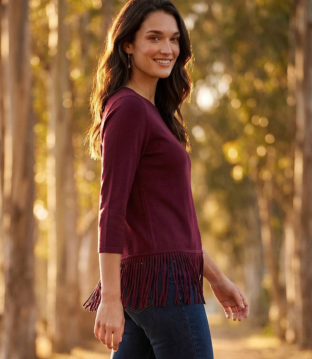 A woman wearing a burgundy three-quarter sleeve top with fringe trim at the hem, paired with dark blue jeans, standing in a sunlit forest of trees.