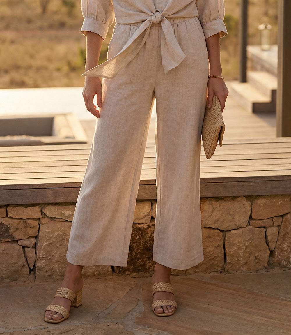 Woman in beige outfit standing outdoors with stone wall and wooden bench in background