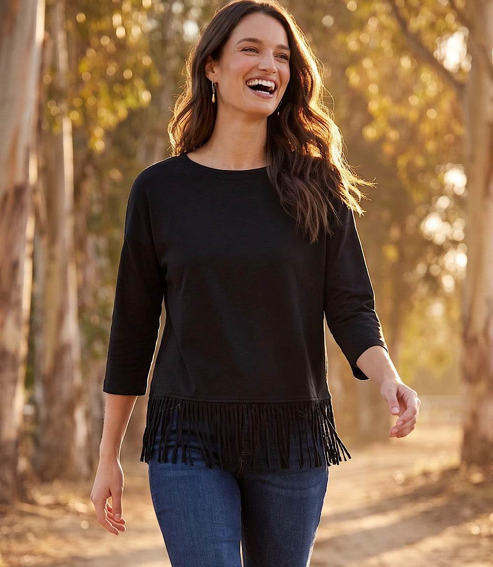 A woman smiling outdoors on a sunlit tree-lined path, wearing a black long-sleeve top with fringe trim at the hem and blue jeans.