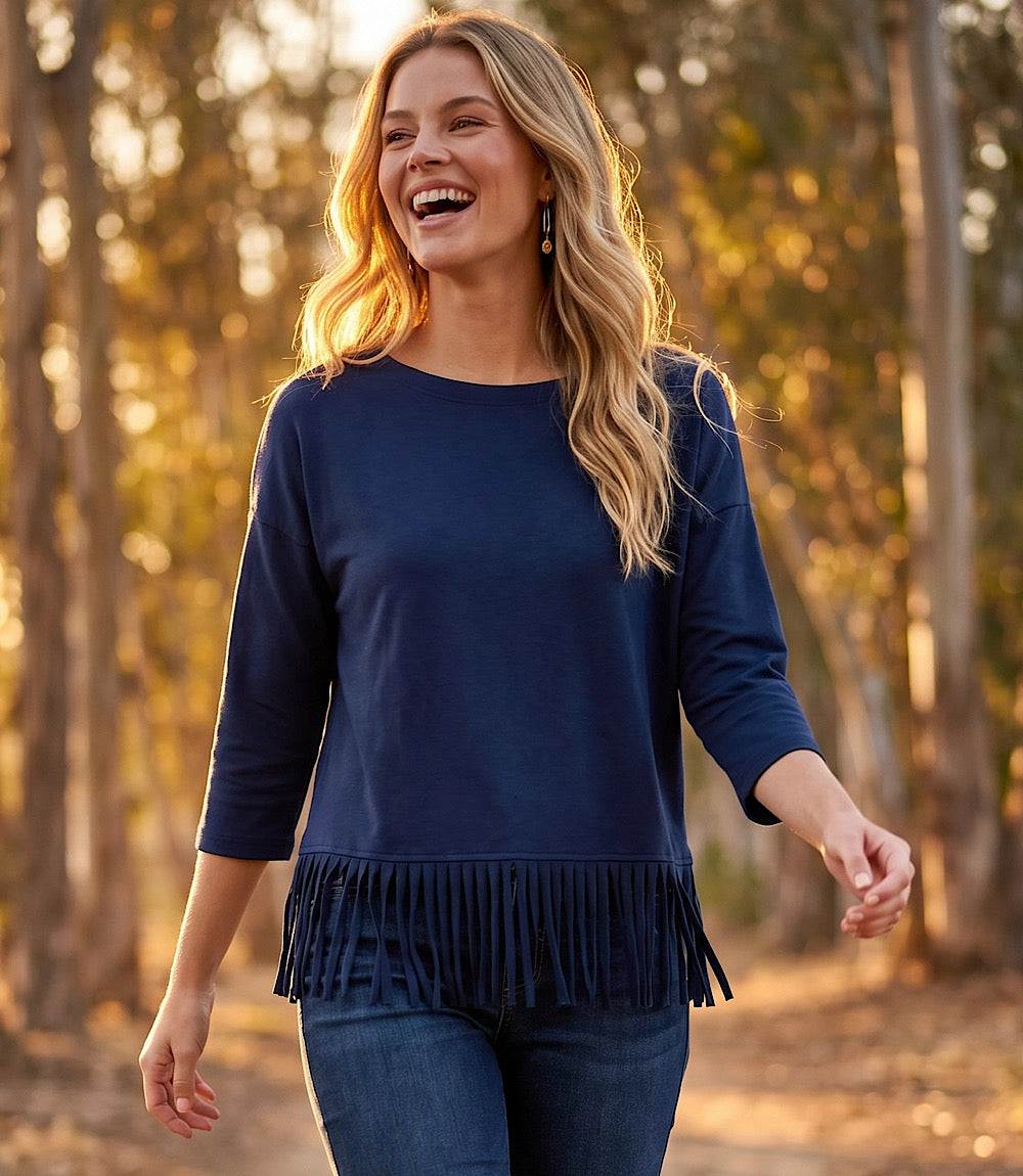 A woman smiling outdoors wearing a navy blue long-sleeve top with fringe trim at the hem, paired with blue jeans, set against a warm, sunlit forest background.