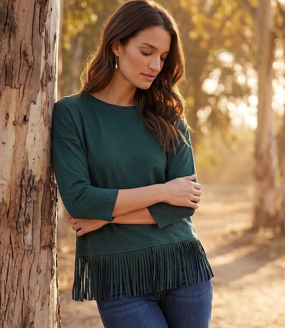 A woman wearing a dark green long-sleeve top with a fringe hem, leaning against a tree in a sunlit outdoor setting.