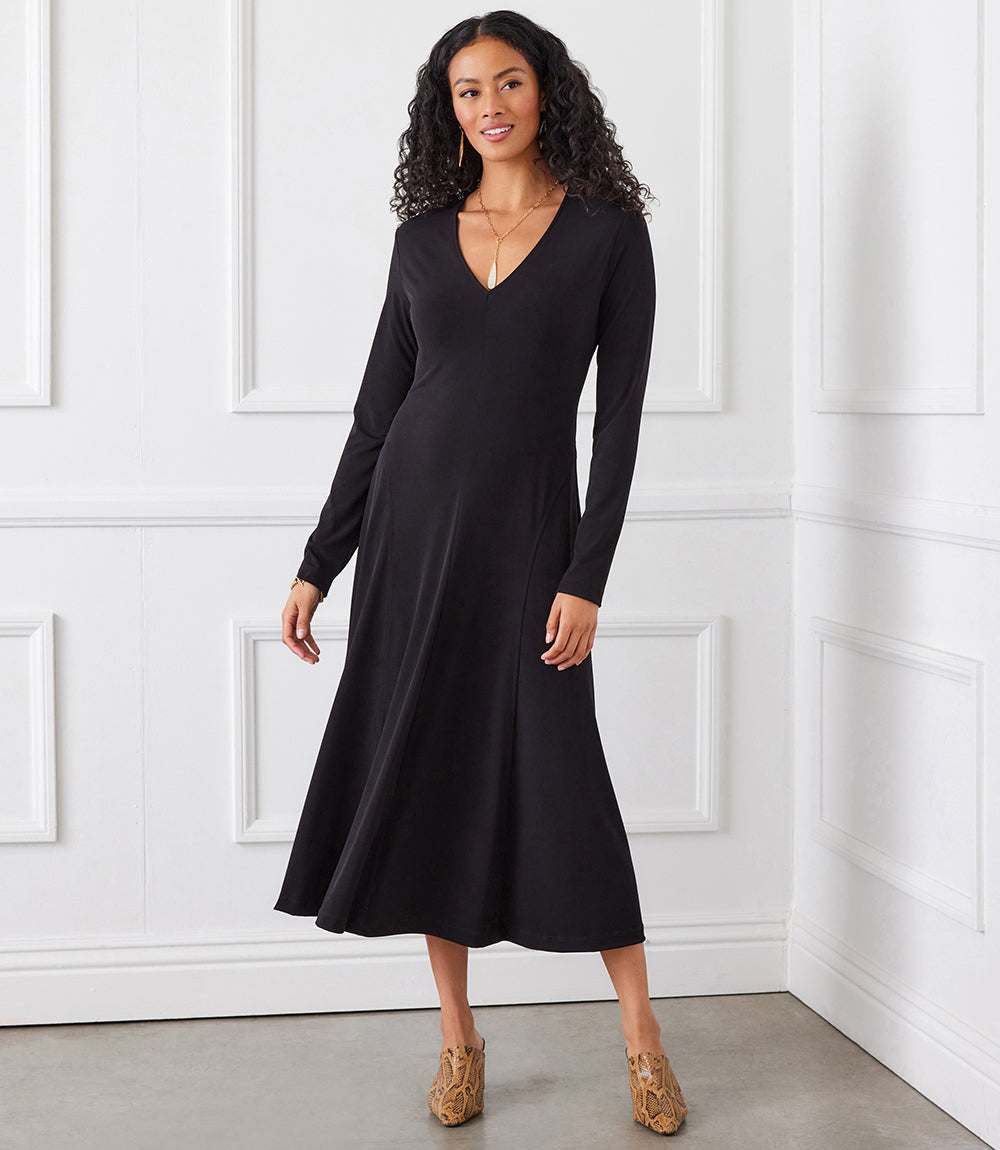 A woman modeling a long-sleeve black midi dress with a V-neckline, standing in front of a white paneled wall.
