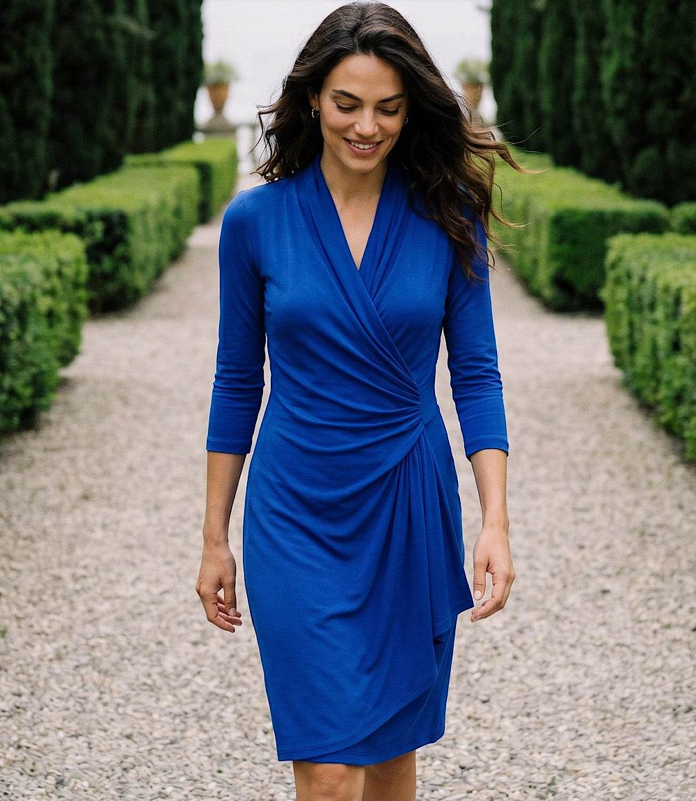 A woman wearing a royal blue wrap-style knee-length dress with three-quarter sleeves walks down a gravel path lined with trimmed hedges.