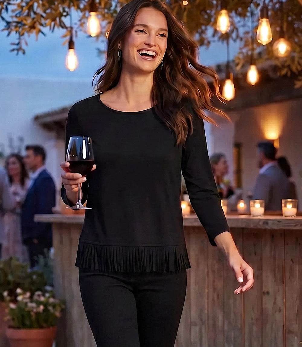 A woman wearing a black long-sleeve top with a fringe hem and black pants walks outdoors at dusk while holding a glass of red wine. The background shows a warm, softly lit patio or bar setting with hanging bulbs and blurred people.