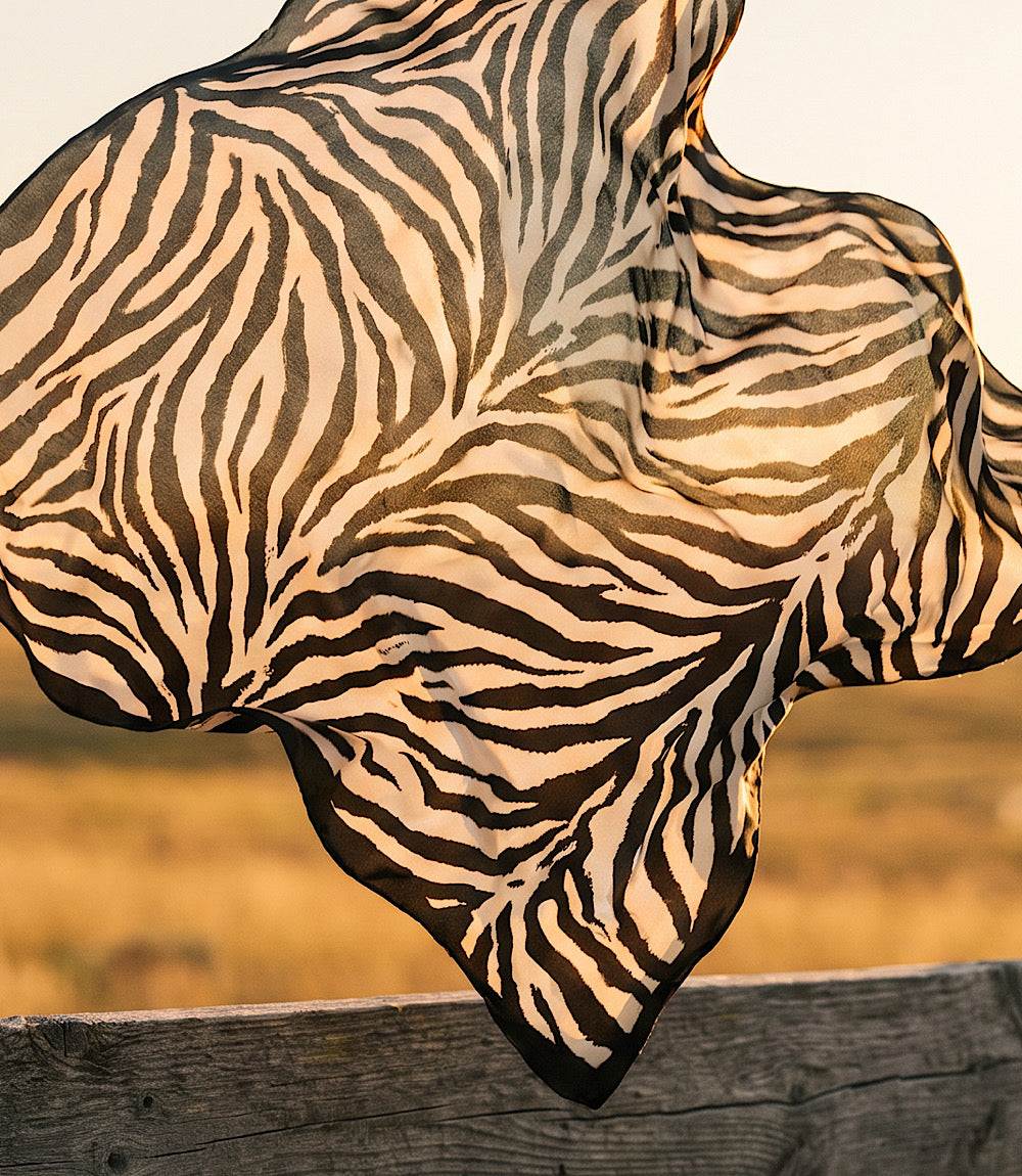 A lightweight square scarf with a zebra print pattern in black and warm beige, draped dynamically outdoors over a rustic wooden fence with a blurred natural background.