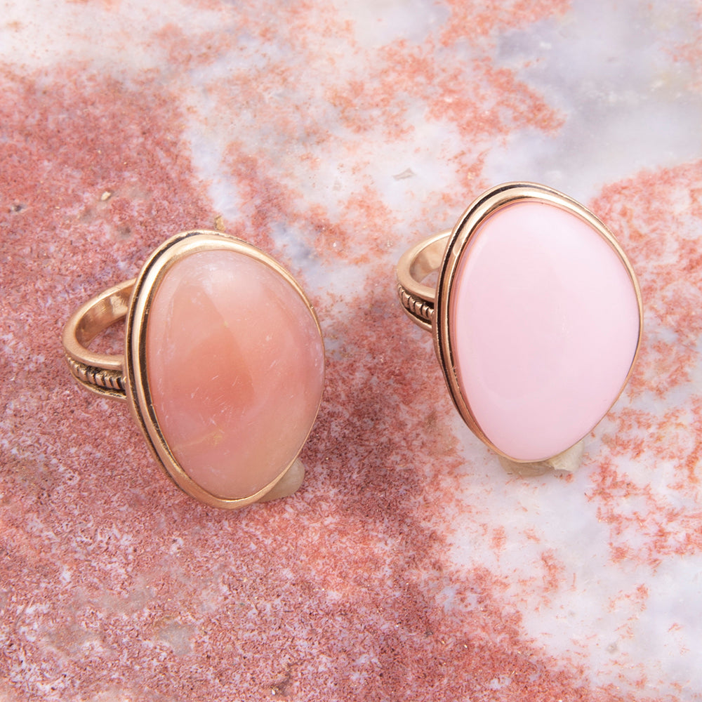 Two oval pink stone rings with gold-tone bezel settings, displayed on a textured pink and white stone surface.