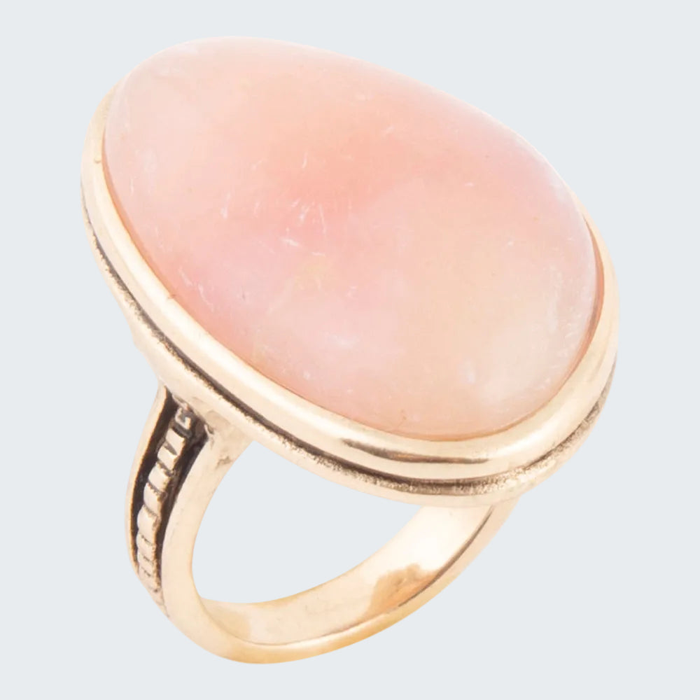 A gold ring featuring a large, smooth, oval-shaped pale pink stone, likely a rose quartz cabochon, set against a plain white background.