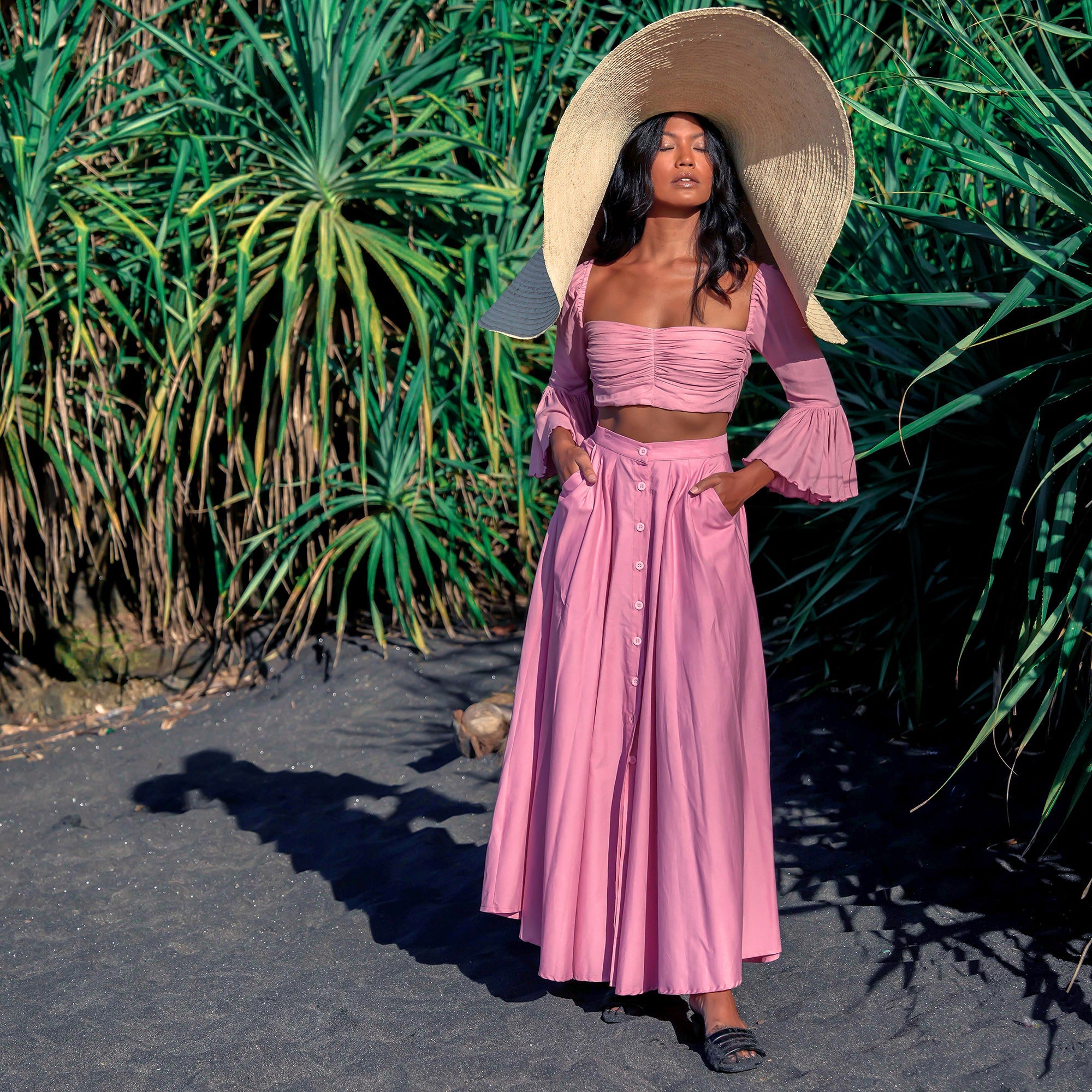 Woman in a pink outfit with a wide-brimmed hat standing in front of green plants.