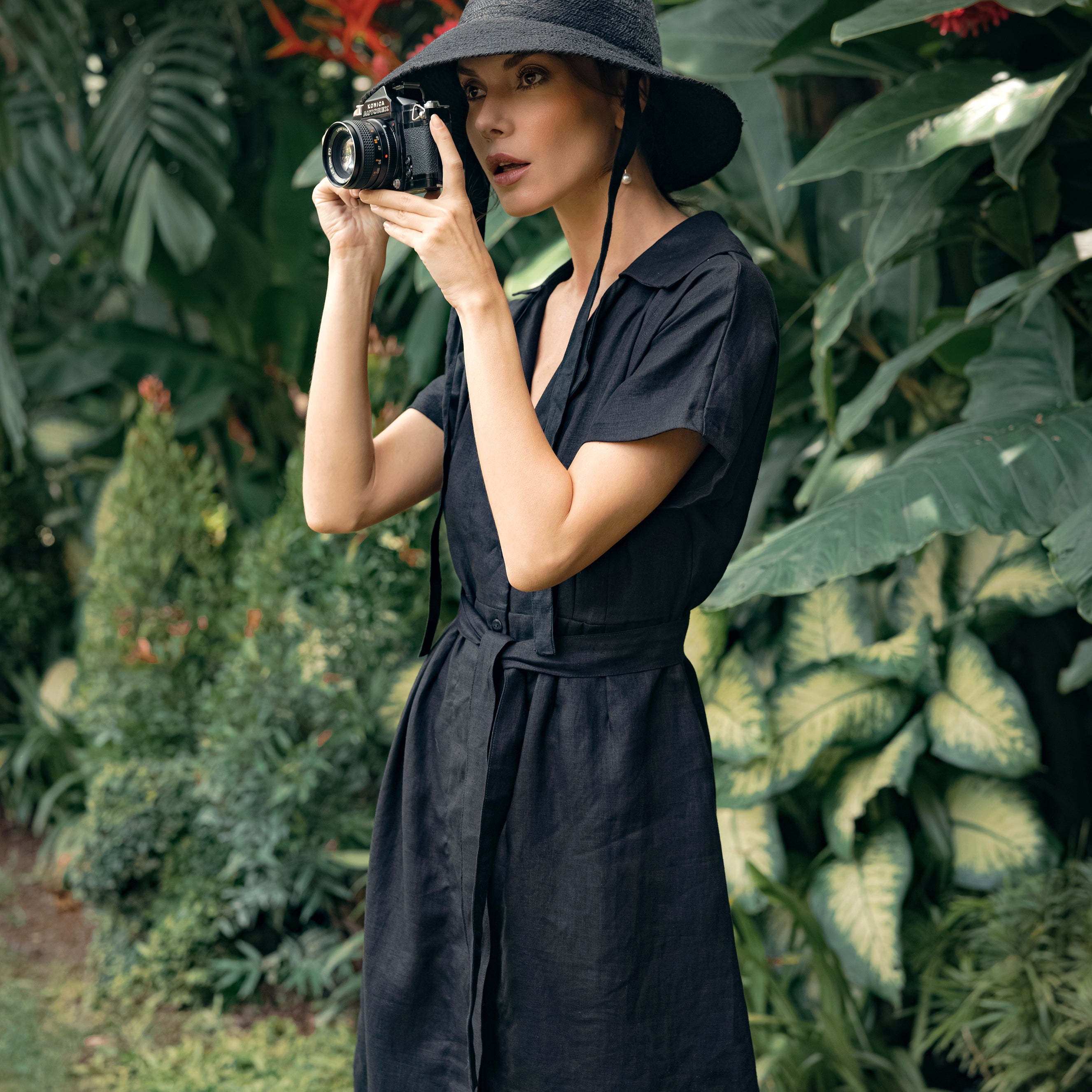 Woman in a black dress and hat holding a camera amidst green foliage