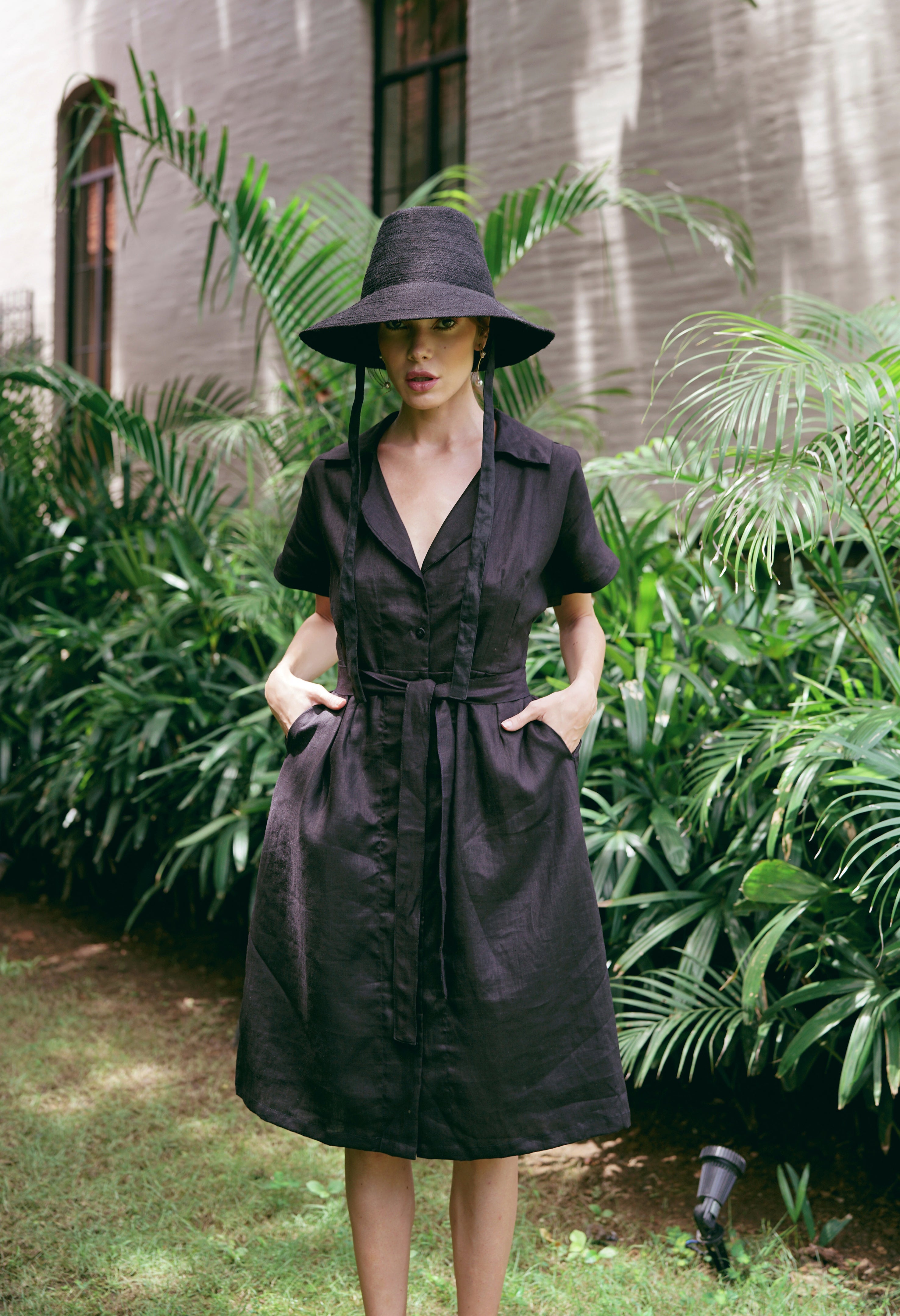 Woman in a black dress and hat standing in a garden with greenery.