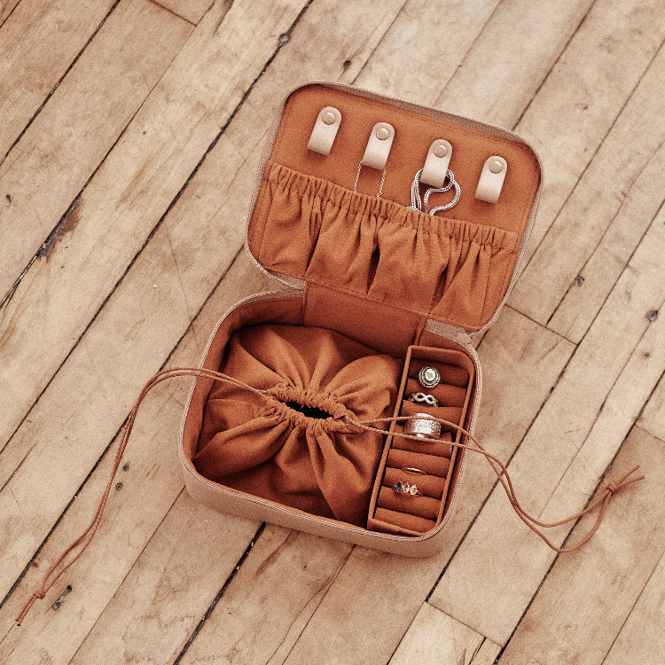 An open tan leather jewelry organizer box on a wooden floor, showing a brown drawstring pouch, ring slots, ring holders, and small hanging loops with rings inside.
