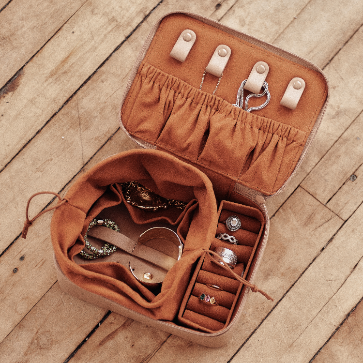 An open tan jewelry organizer case with a suede-like interior, shown on a wooden floor. The case has elastic loops, a drawstring pouch, ring slots, and hanging pegs, with various rings and bracelets displayed inside.