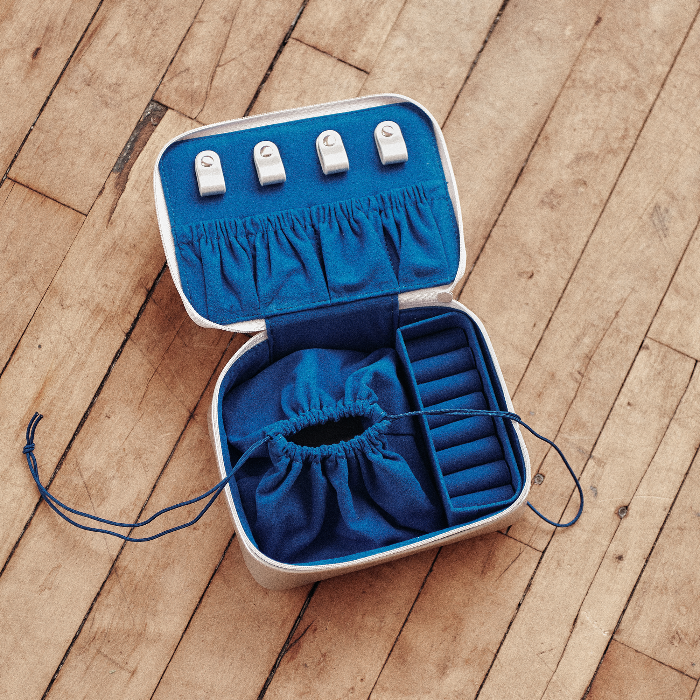 An open jewelry organizer case with a white exterior and bright blue interior, placed on a wooden floor. The case includes four white ring holders on the inside lid, a blue drawstring pouch, and a compartmentalized tray on the bottom right.