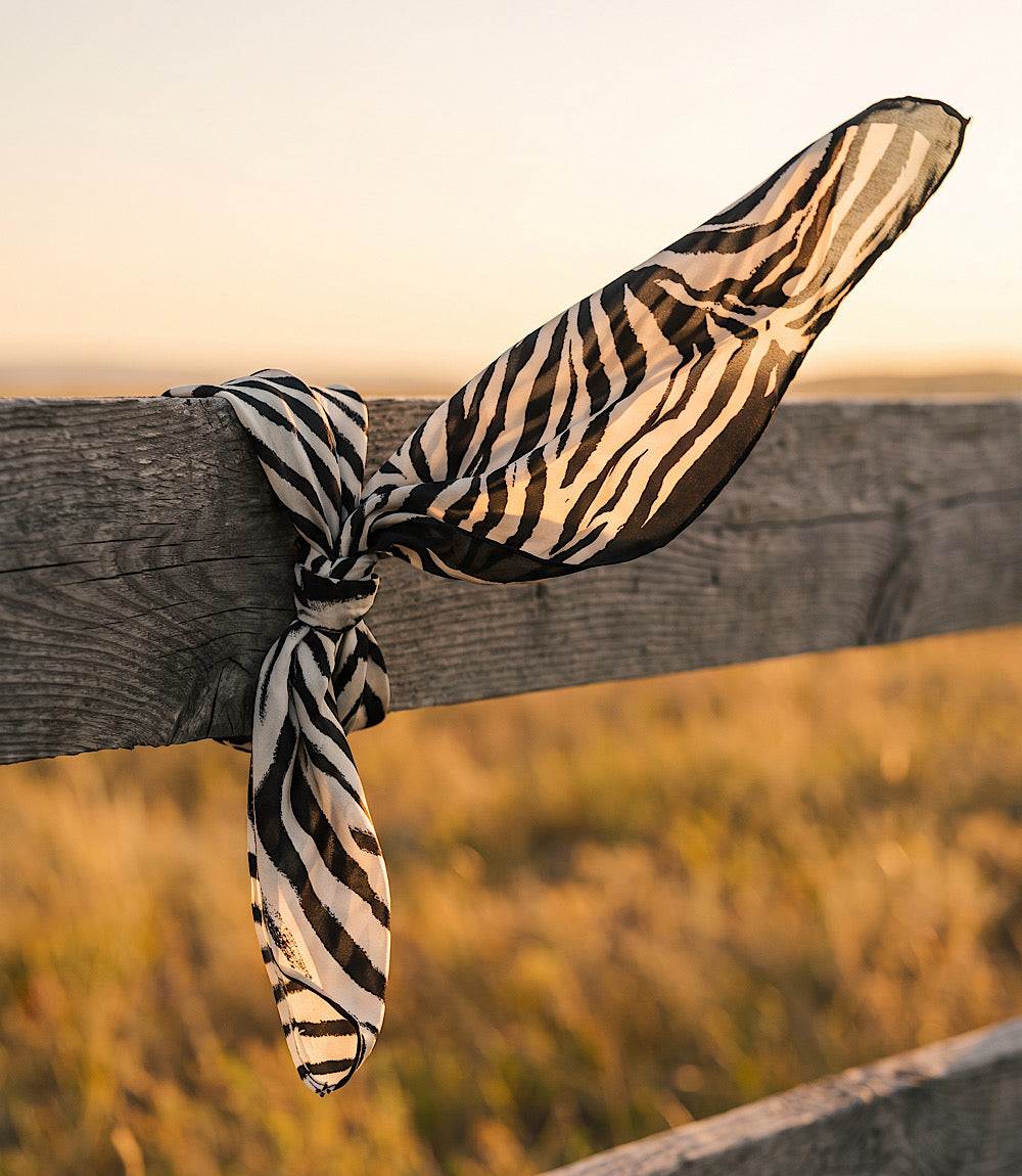 A lightweight scarf with a black and beige zebra stripe pattern tied in a knot and draped over a rustic wooden fence post in an outdoor field at sunset.