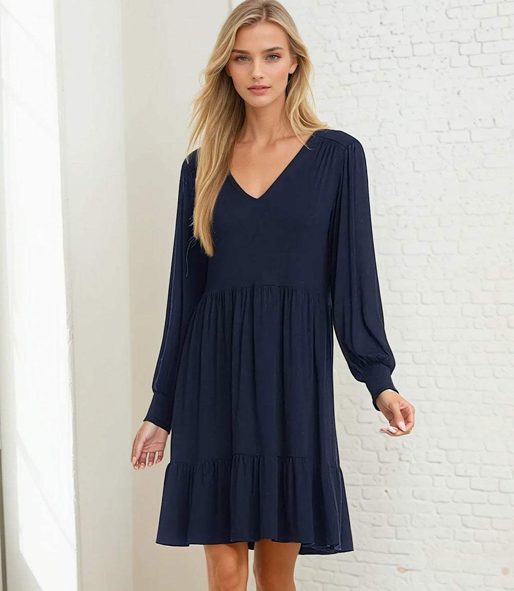 A woman modeling a navy blue long-sleeve V-neck dress with a tiered skirt, standing against a white brick wall.