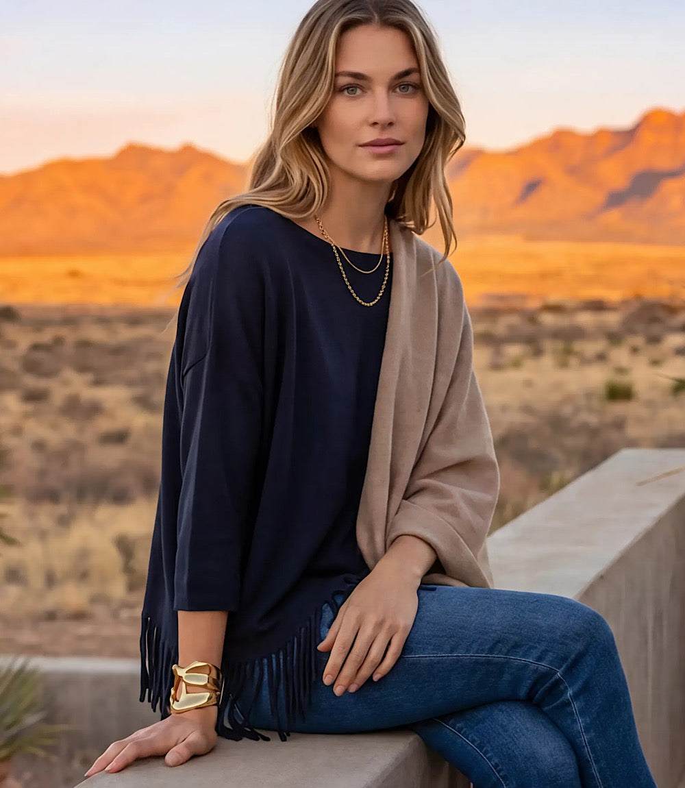 A woman sits on a concrete ledge in a desert landscape at sunset, wearing a navy blue top with fringe trim, a beige shawl draped over her shoulders, blue jeans, and gold jewelry.