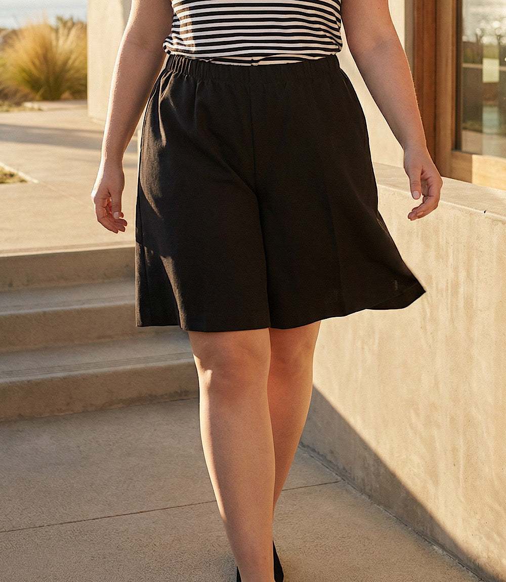 A person wearing black wide-leg shorts and a black-and-white striped top, standing outdoors near a beige wall and steps in warm sunlight.