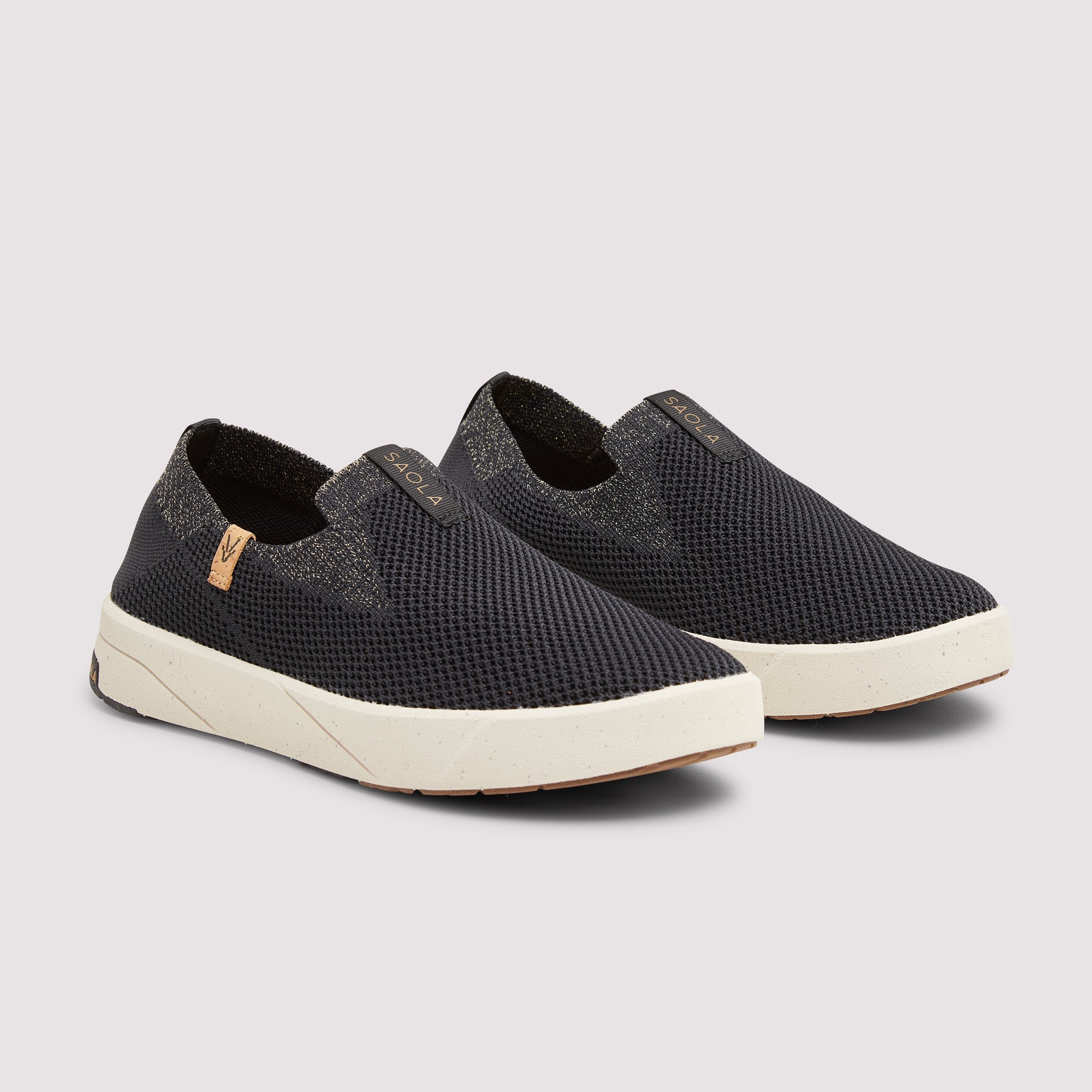 A pair of slip-on mesh sneakers in black with textured gray heel panels and thick off-white soles, shown on a light gray background.