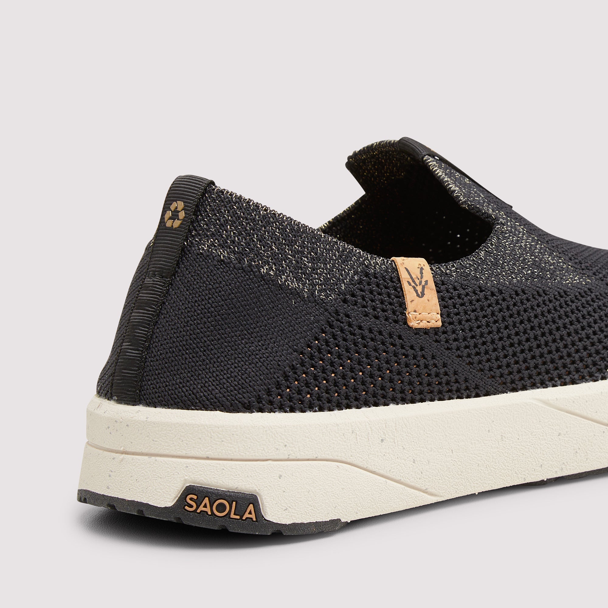A close-up of a black slip-on mesh sneaker by SAOLA, showing the textured knit upper, a small tan leather logo tab, and a chunky off-white sole.