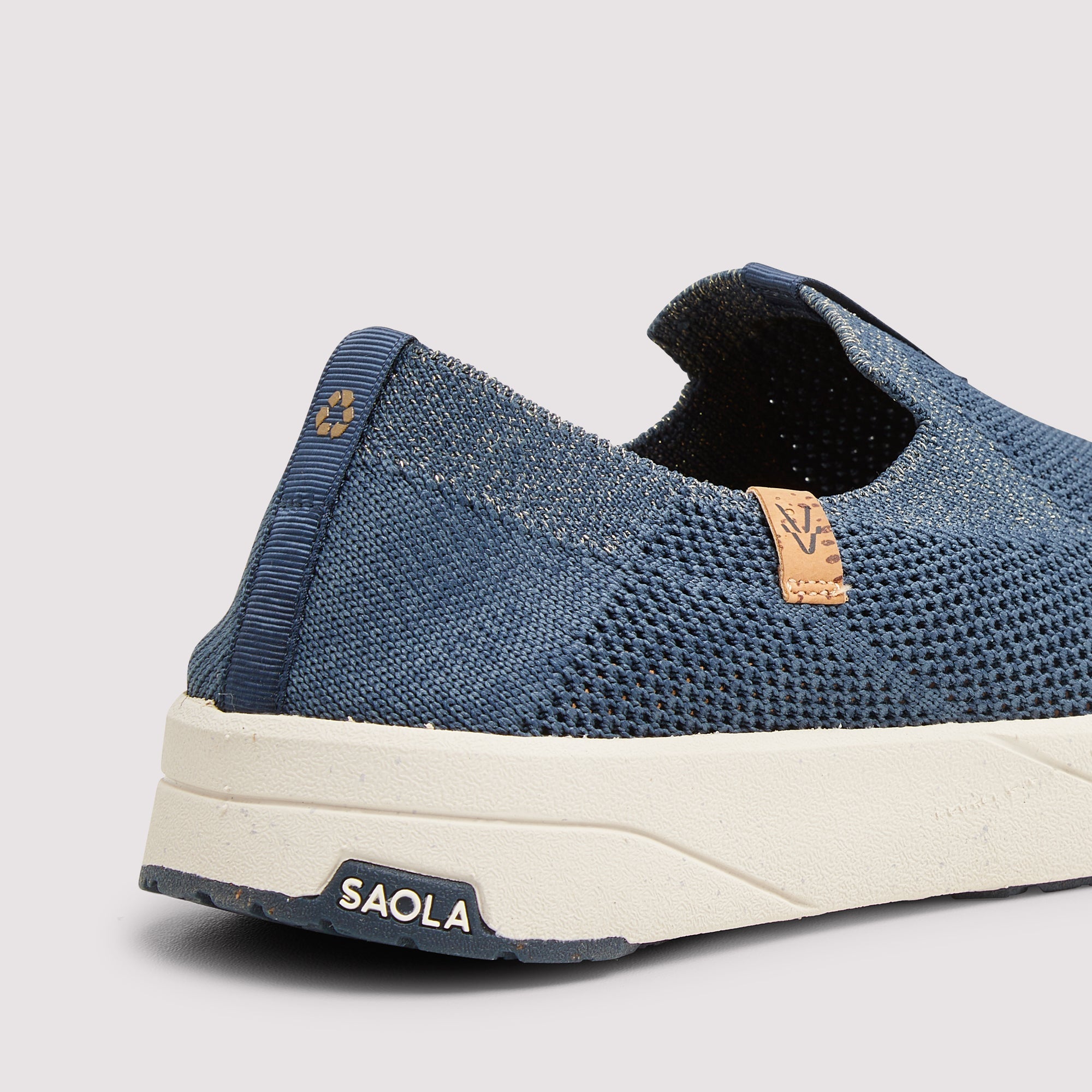 A close-up shot of a blue knit slip-on shoe by Saola, featuring a textured knit upper, a cream-colored midsole, and a small tan leather brand tag on the side.