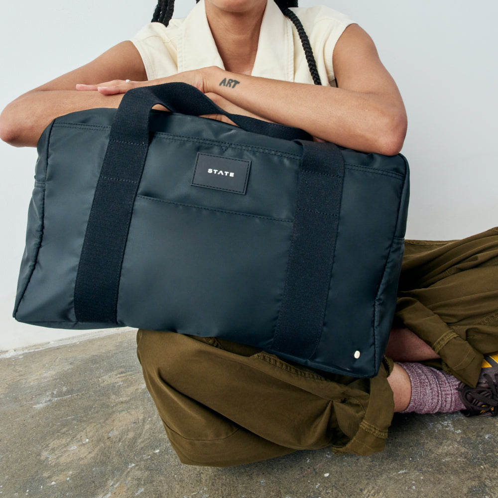 Person holding a navy blue bag with 'STAUD' label on a concrete floor.