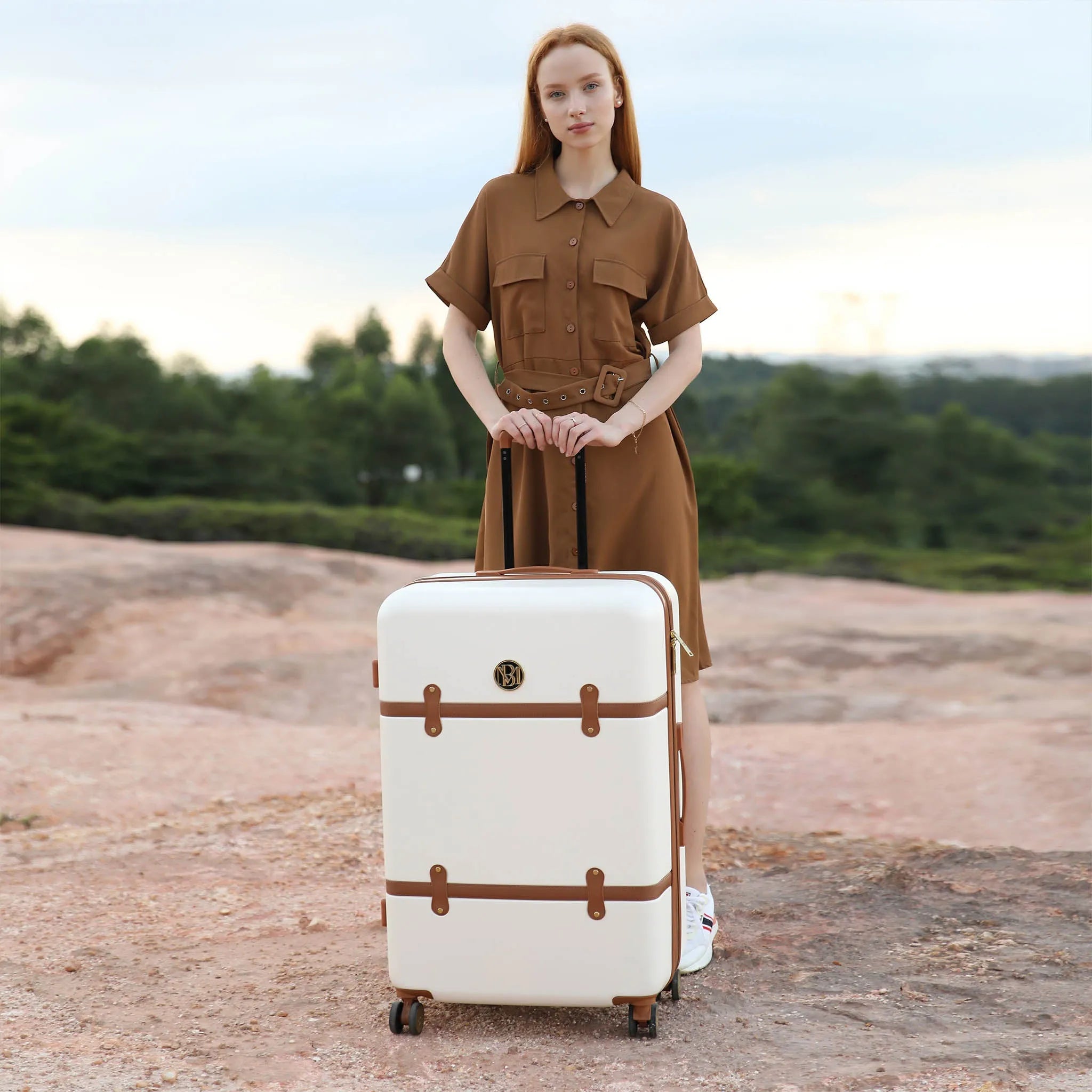 Woman in a brown dress standing next to a white suitcase with brown straps in an outdoor setting.