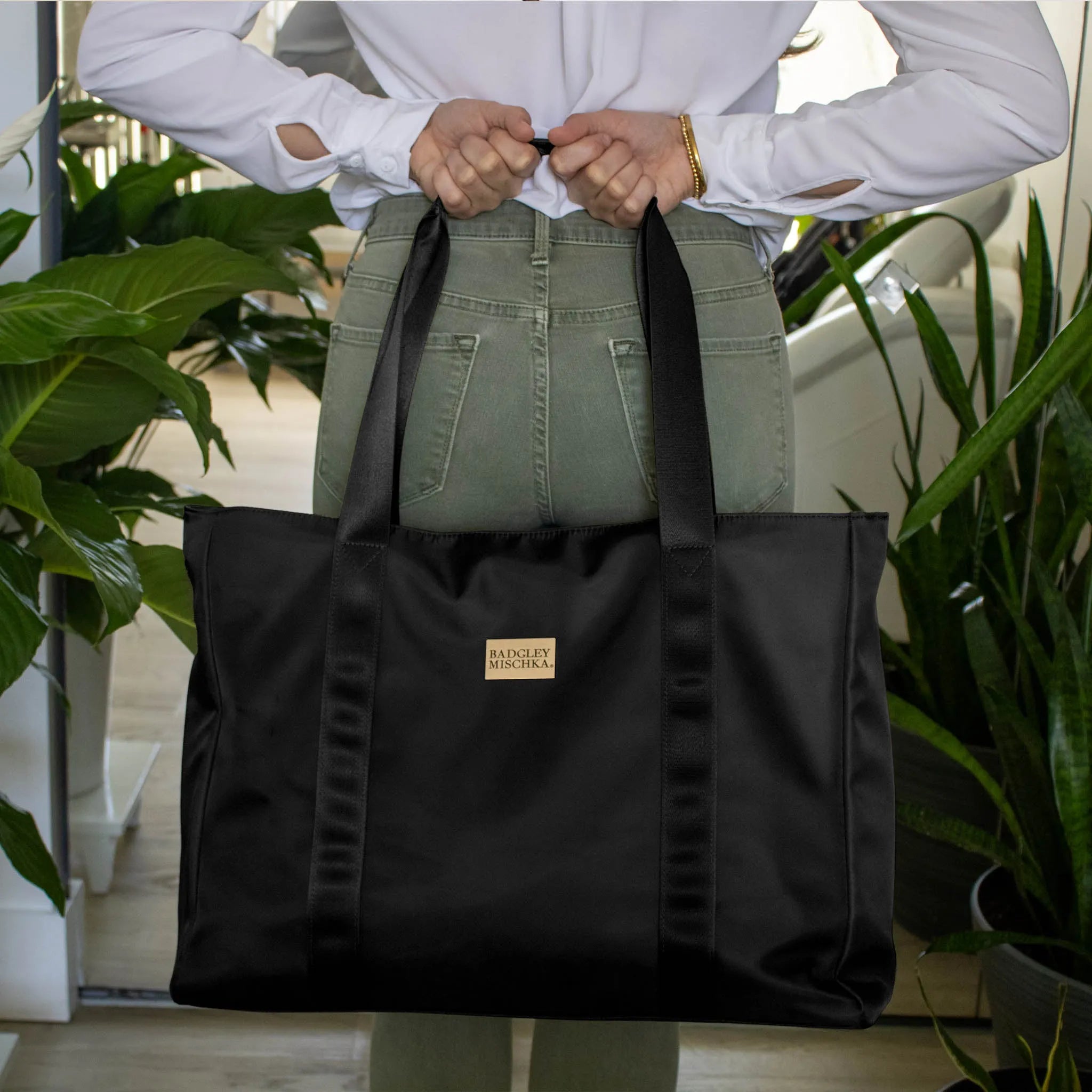Person holding a black tote bag with a visible brand logo, surrounded by plants.