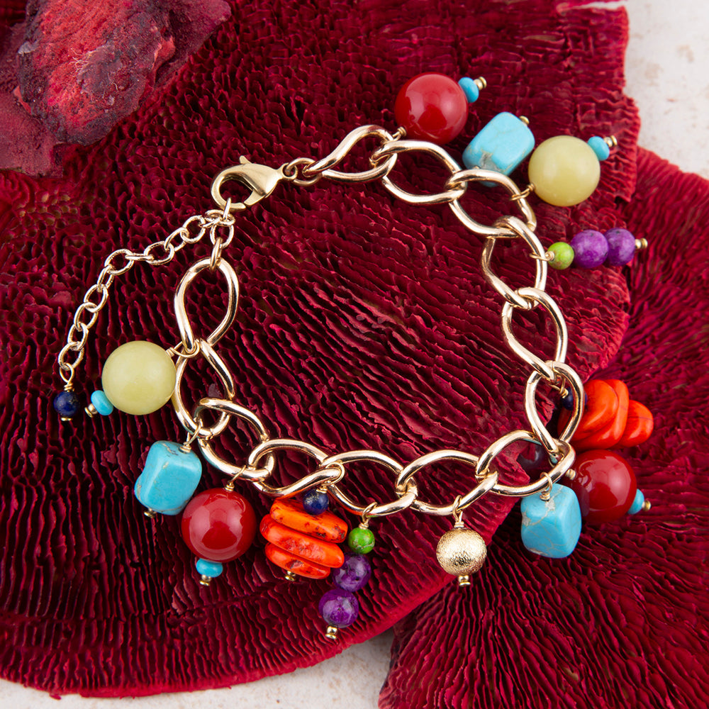 A gold-tone chain bracelet with colorful charm beads and pendants, displayed on a textured deep red fabric background.