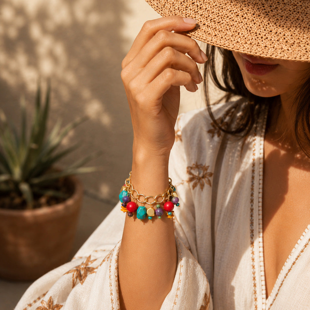 A close-up lifestyle shot of a woman wearing a delicate gold chain bracelet adorned with colorful, round bead charms. She is wearing a white embroidered bohemian-style top and a woven straw hat, with a potted succulent in the background under warm sunlight.