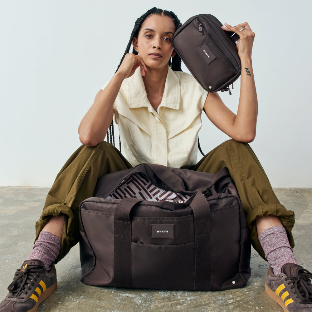 Woman sitting on the floor with a large brown bag in front of her, holding a smaller bag above her head.
