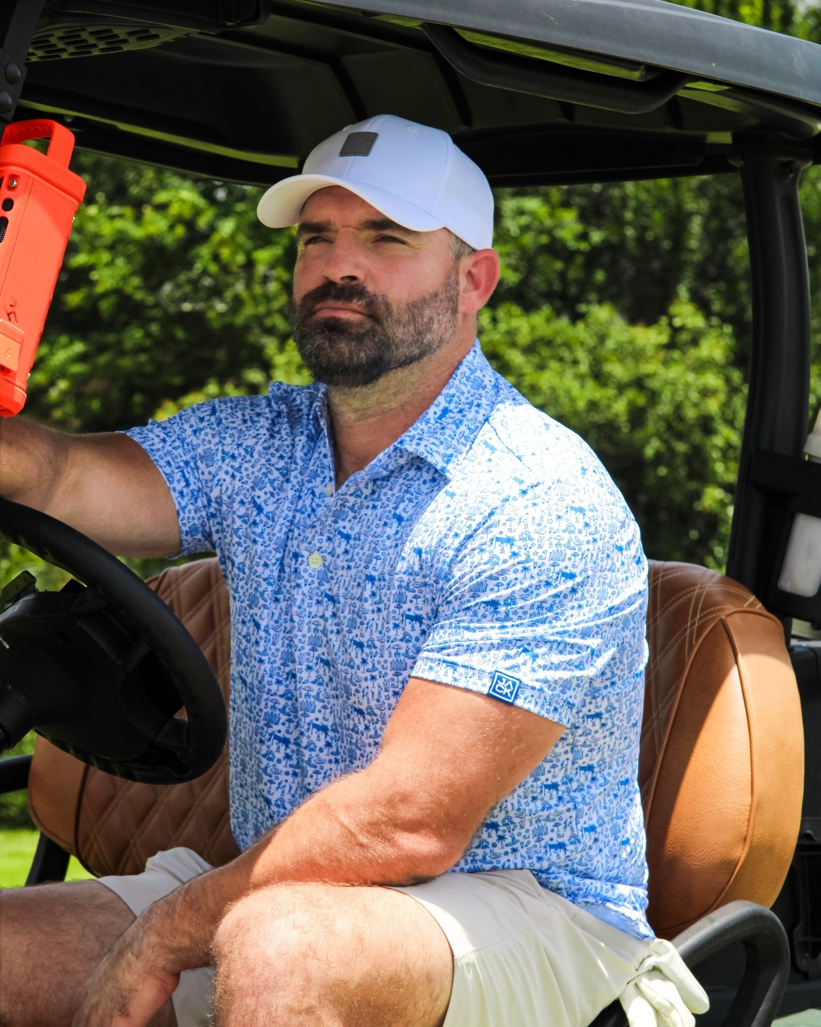 A man with a beard wearing a blue patterned short-sleeve button-up shirt, white shorts, and a white baseball cap sits inside a golf cart on a green course.